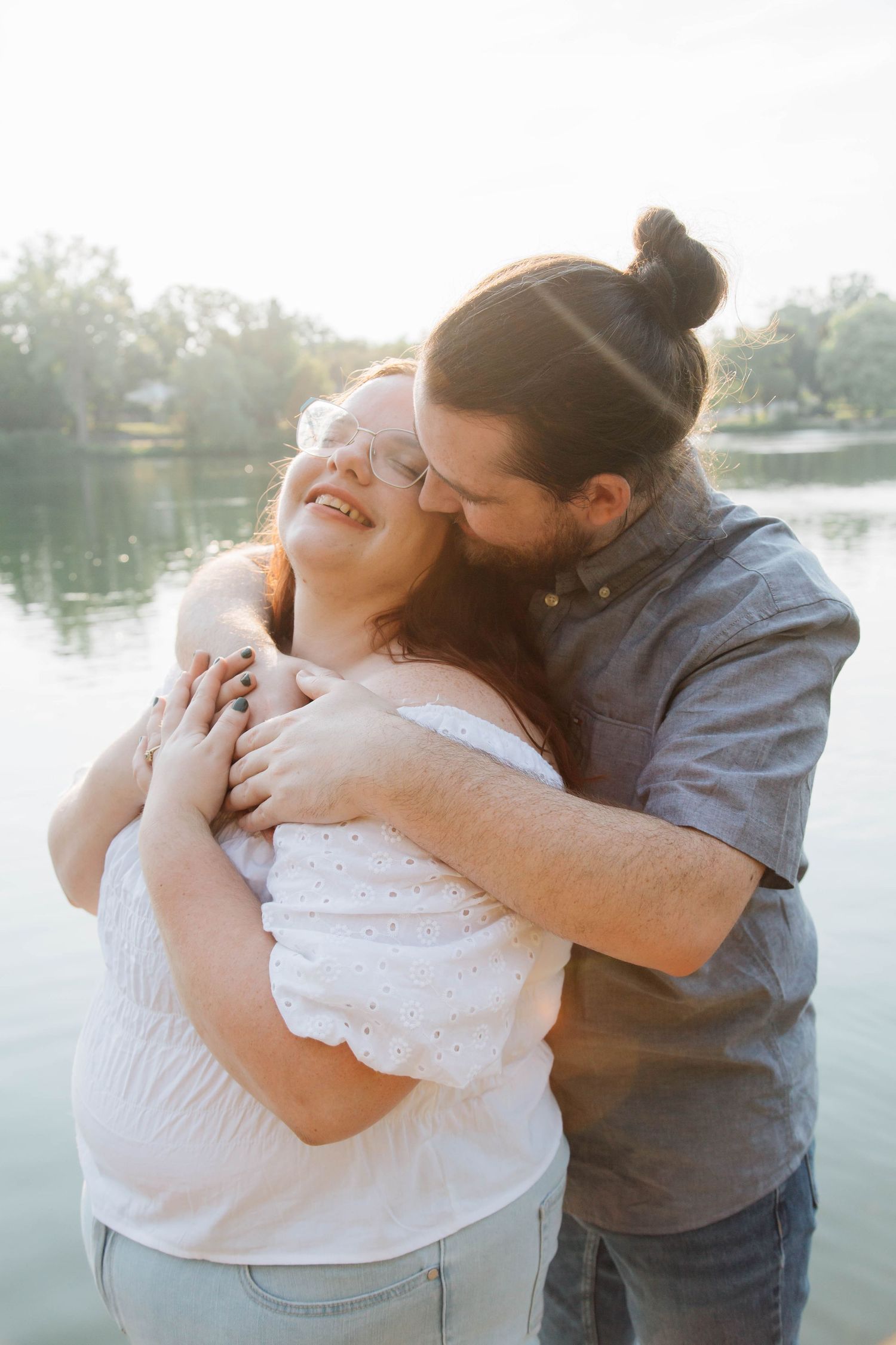 Man tenderly embracing and kissing a laughing woman by a sunlit lake in warm golden light.