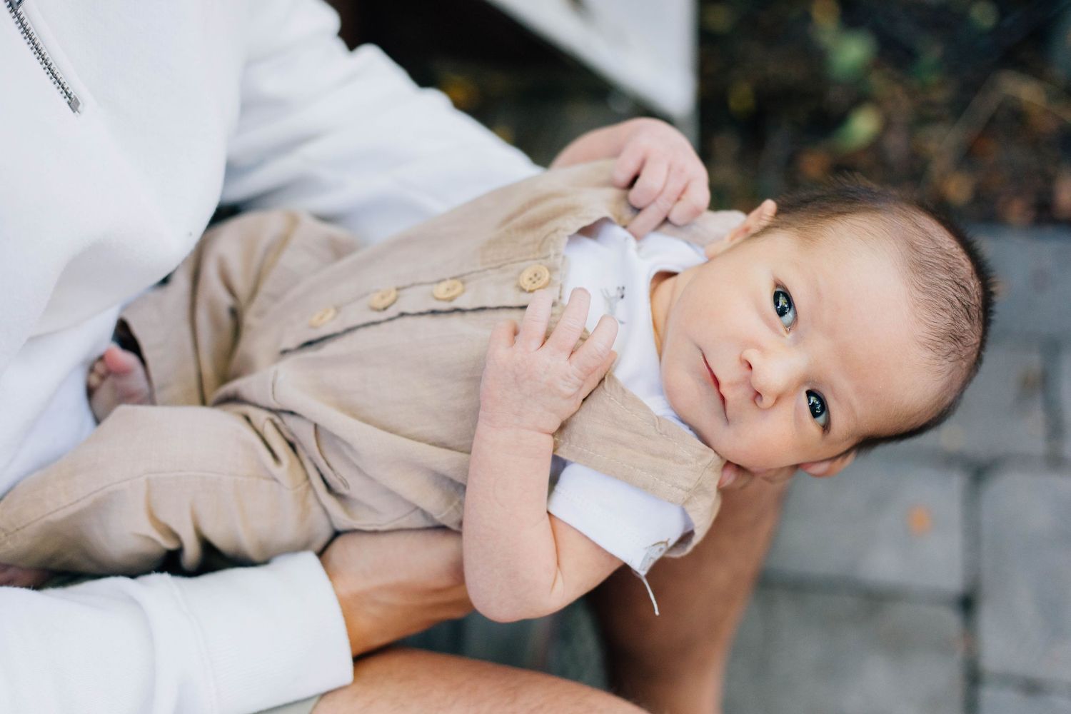 Close-up of wide-eyed newborn baby in tan linen romper being cradled in parents arms outdoors.
