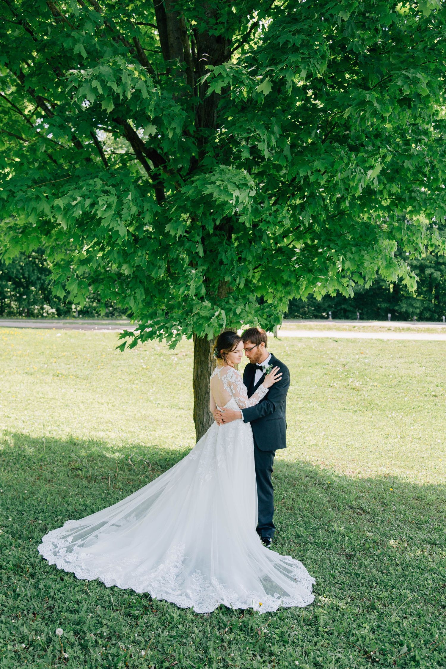 Bride and groom embracing under a large green tree in a sunlit park setting.