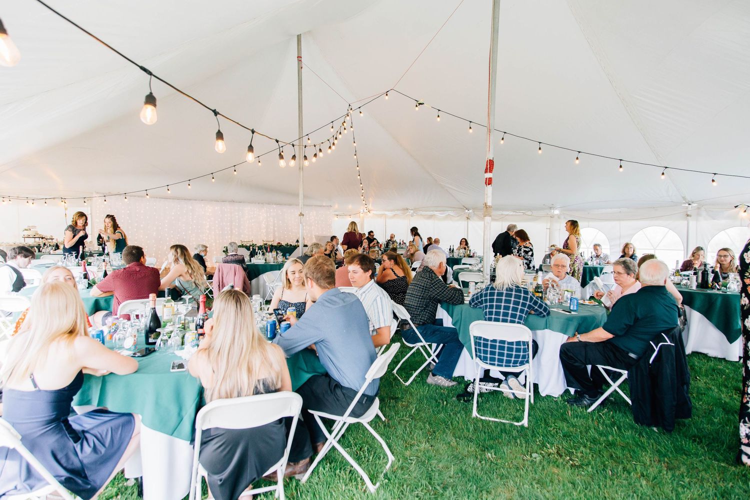 Guests seated at round tables with green linens enjoy a wedding reception under a white tent with string lights.