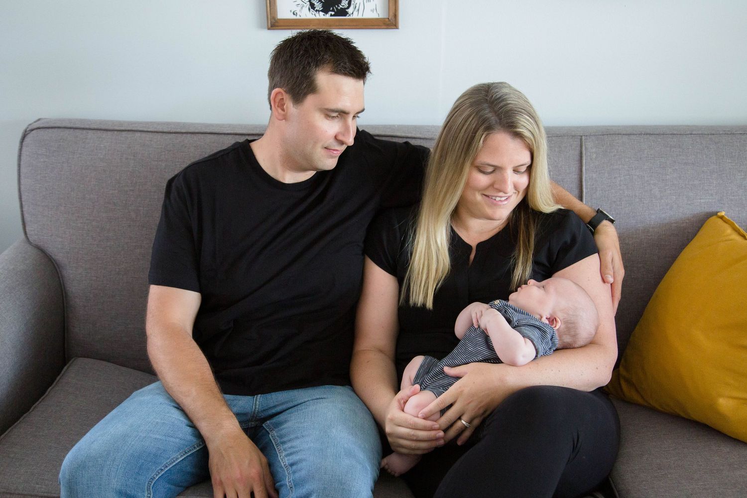 Couple in black shirts admiring their sleeping newborn baby on gray couch at home.
