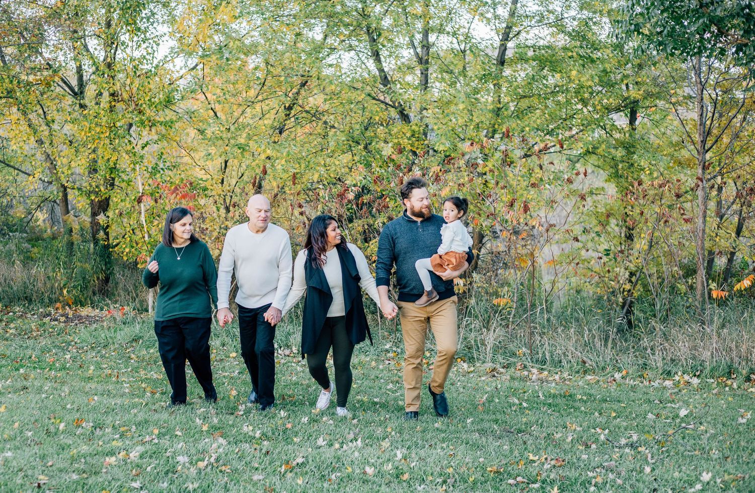 Extended family of five walking together through a park with vibrant autumn foliage trees.