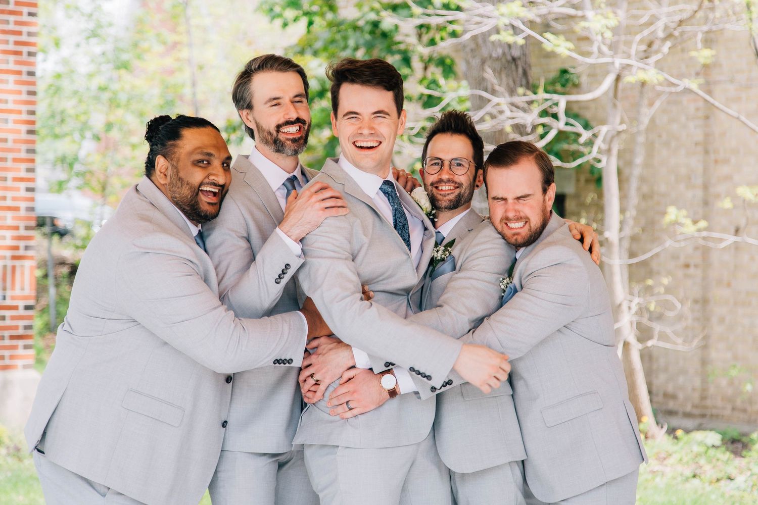 Groom laughs as groomsmen in matching gray suits huddle around him for fun wedding party photo.