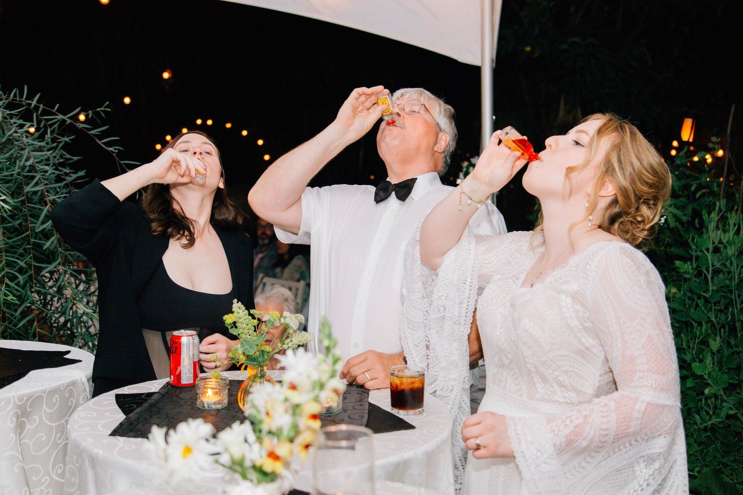 Three people taking celebratory shots at a wedding reception with floral centerpiece table.