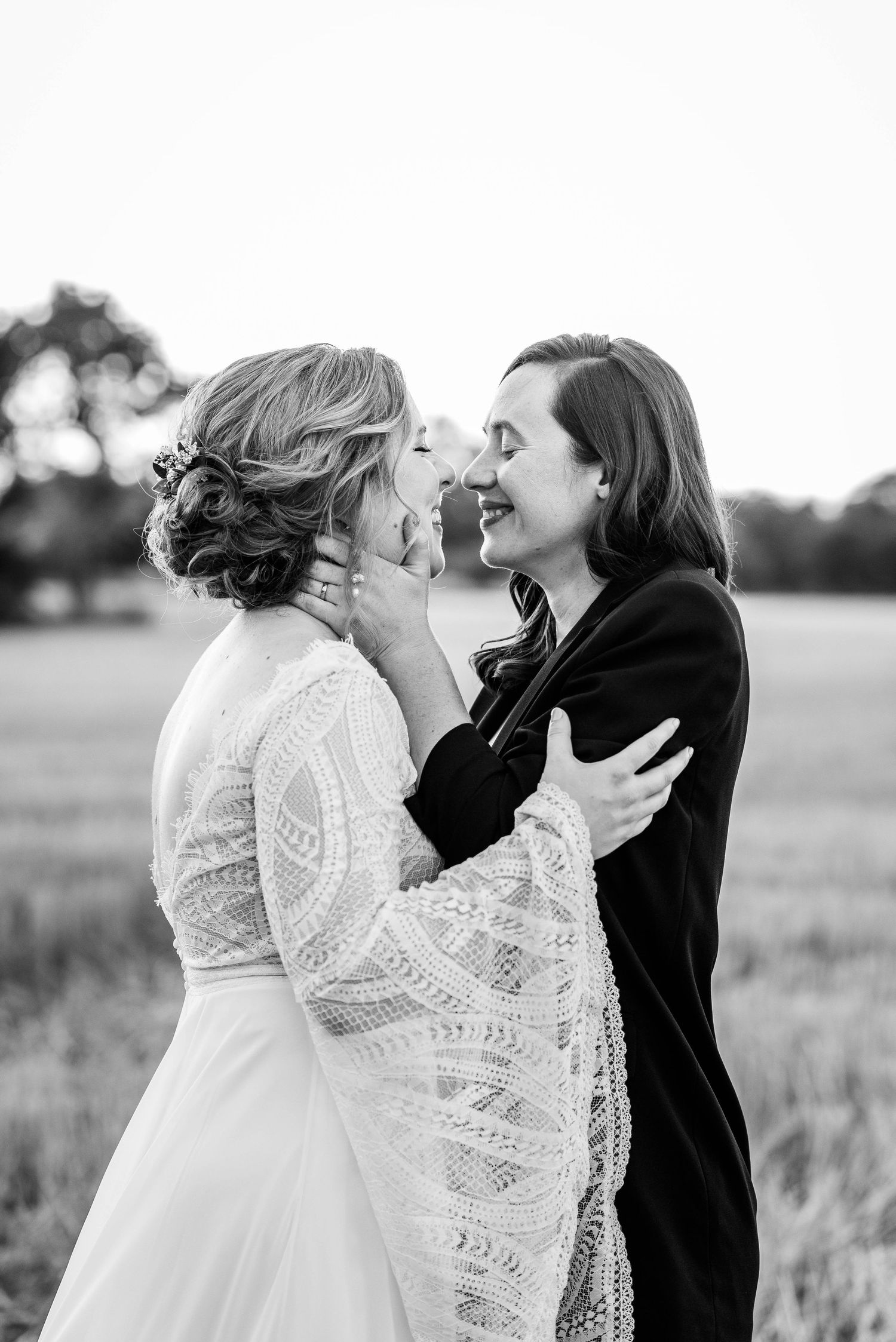 Black and white photo of two brides laughing together in open field, one in lace gown, one in black suit.