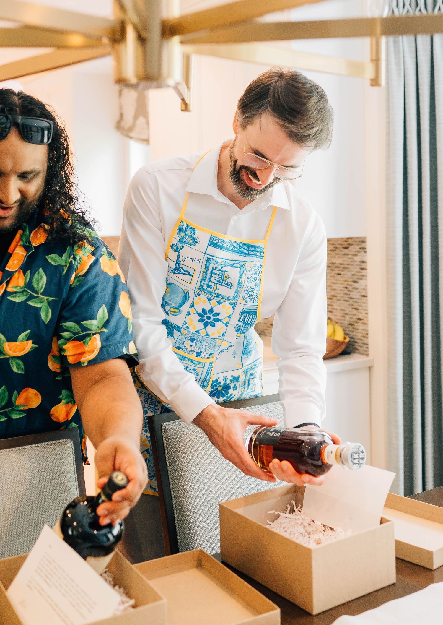 Two men smile while assembling wedding gift boxes with bottles in a bright kitchen setting.
