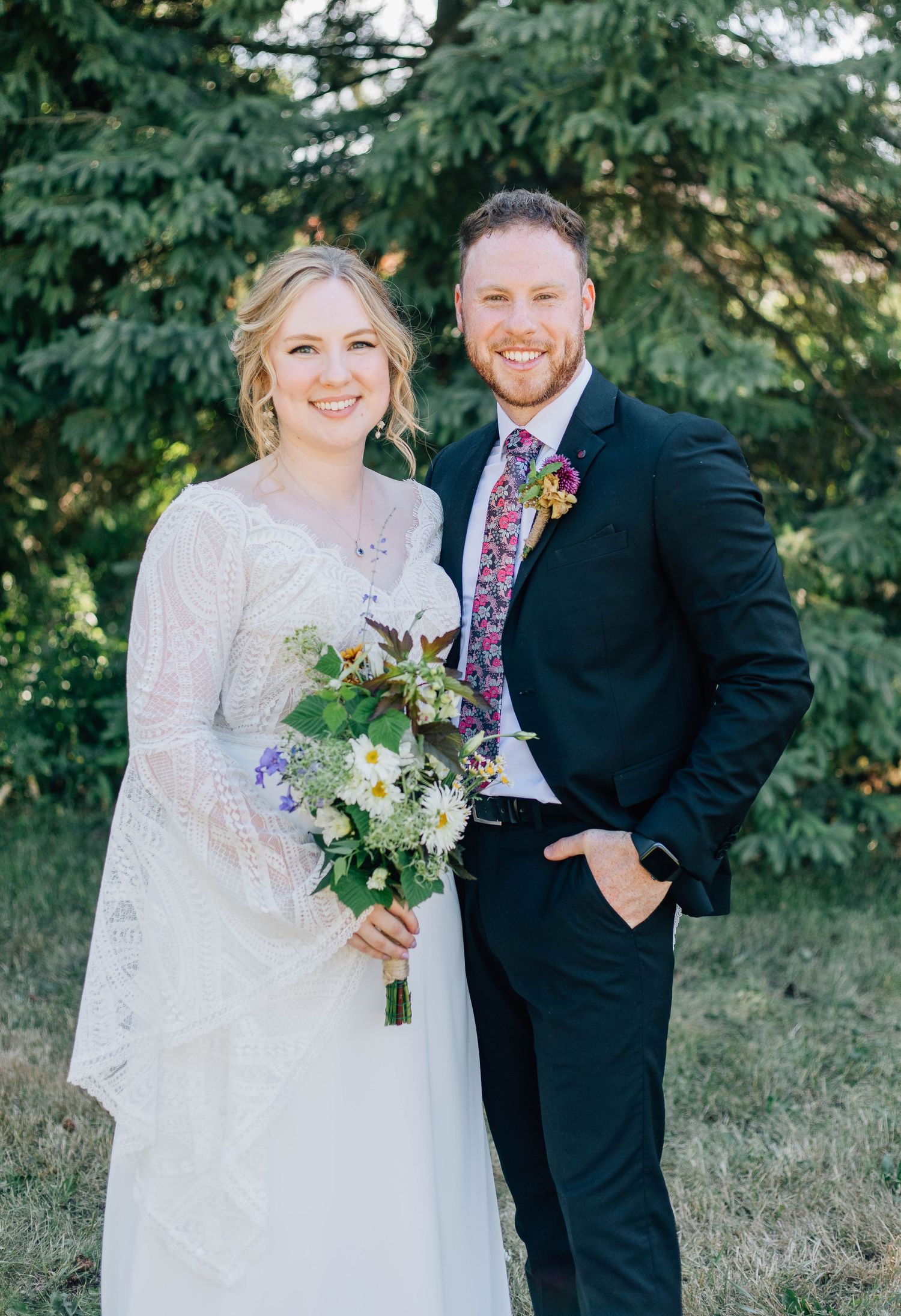 Smiling bride in lace gown holds wildflower bouquet beside groom in dark suit with floral tie outdoors.