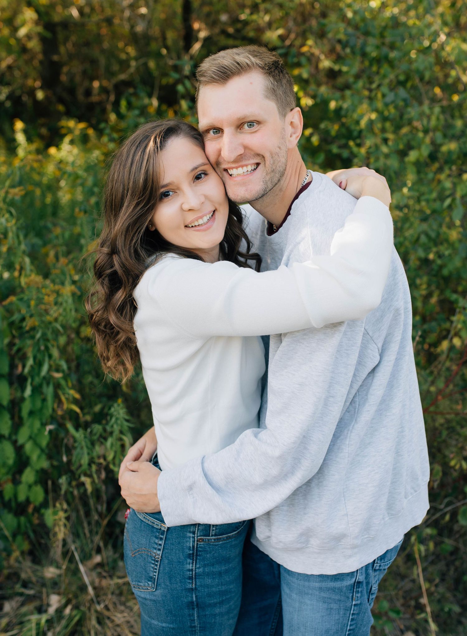 Smiling couple hugging outdoors with lush green foliage in the background.