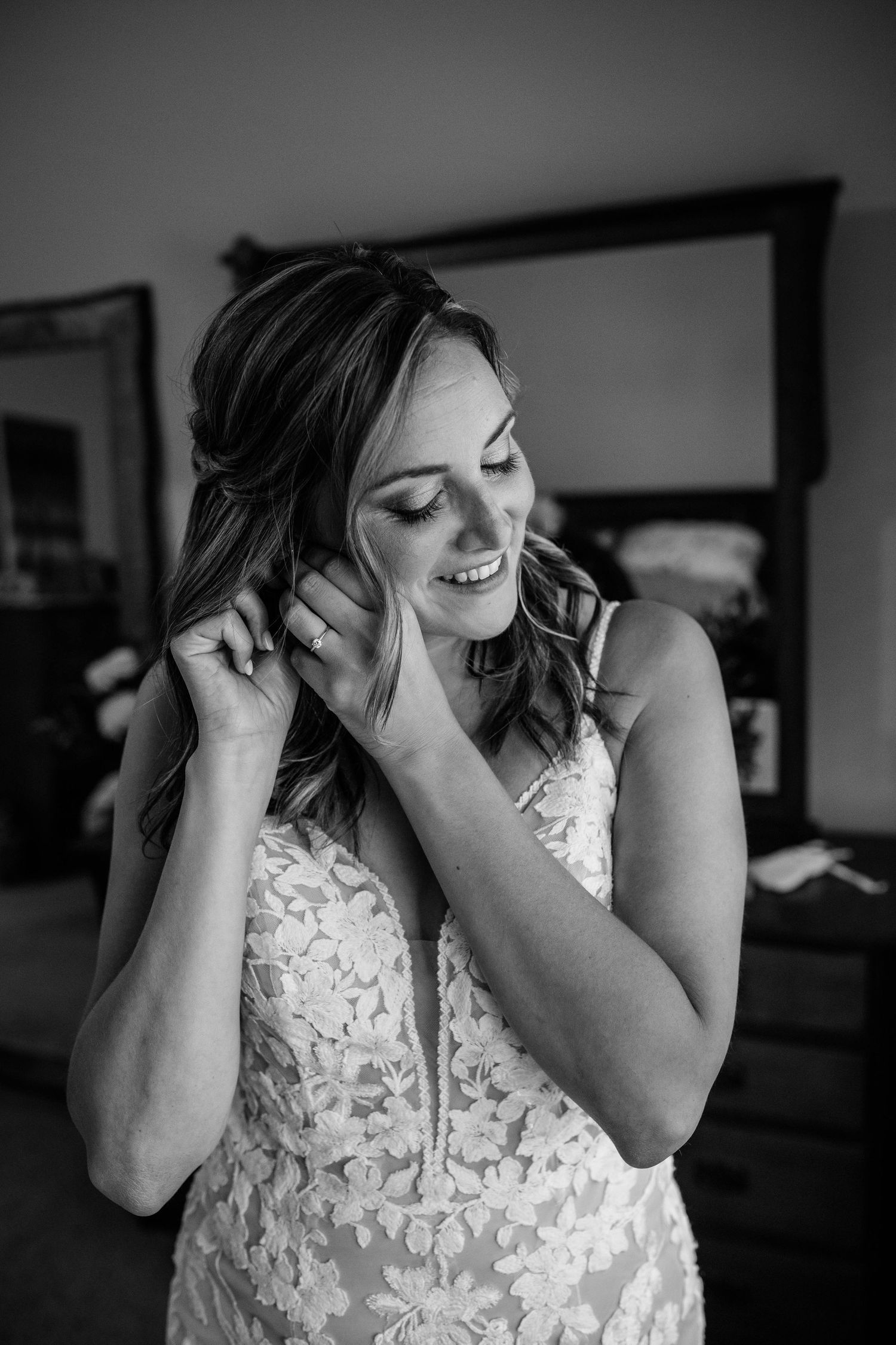 Black and white portrait of a smiling bride in lace gown putting on earrings before her wedding.