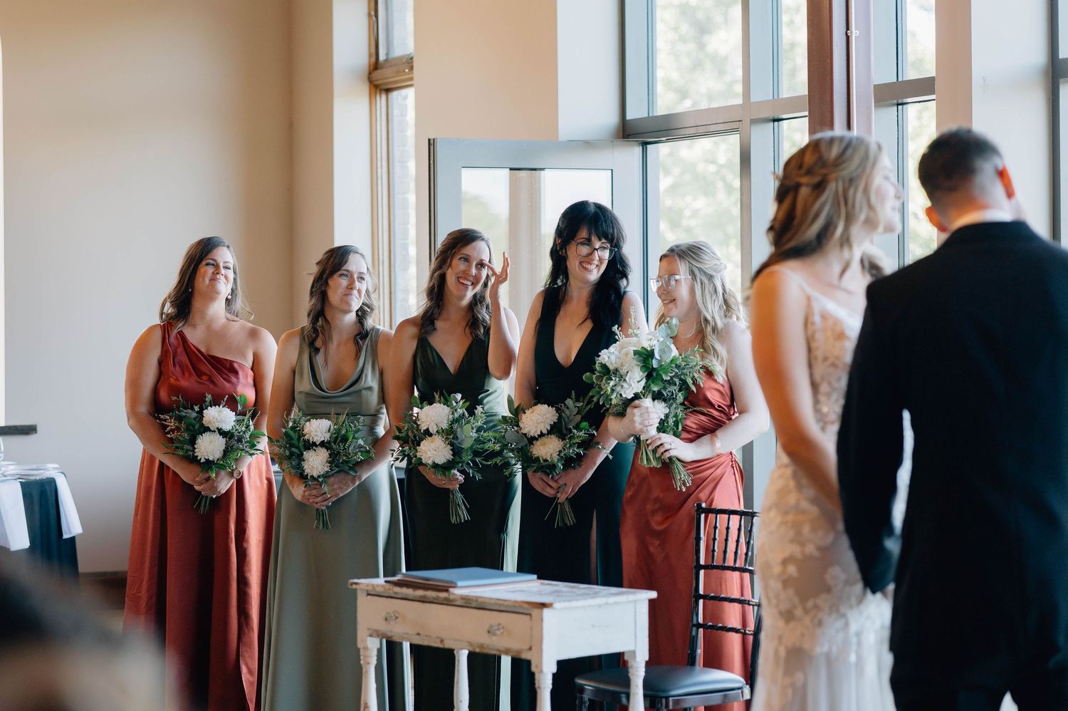 Bridesmaids in mixed jewel-tone dresses holding white bouquets smiling at bride.
