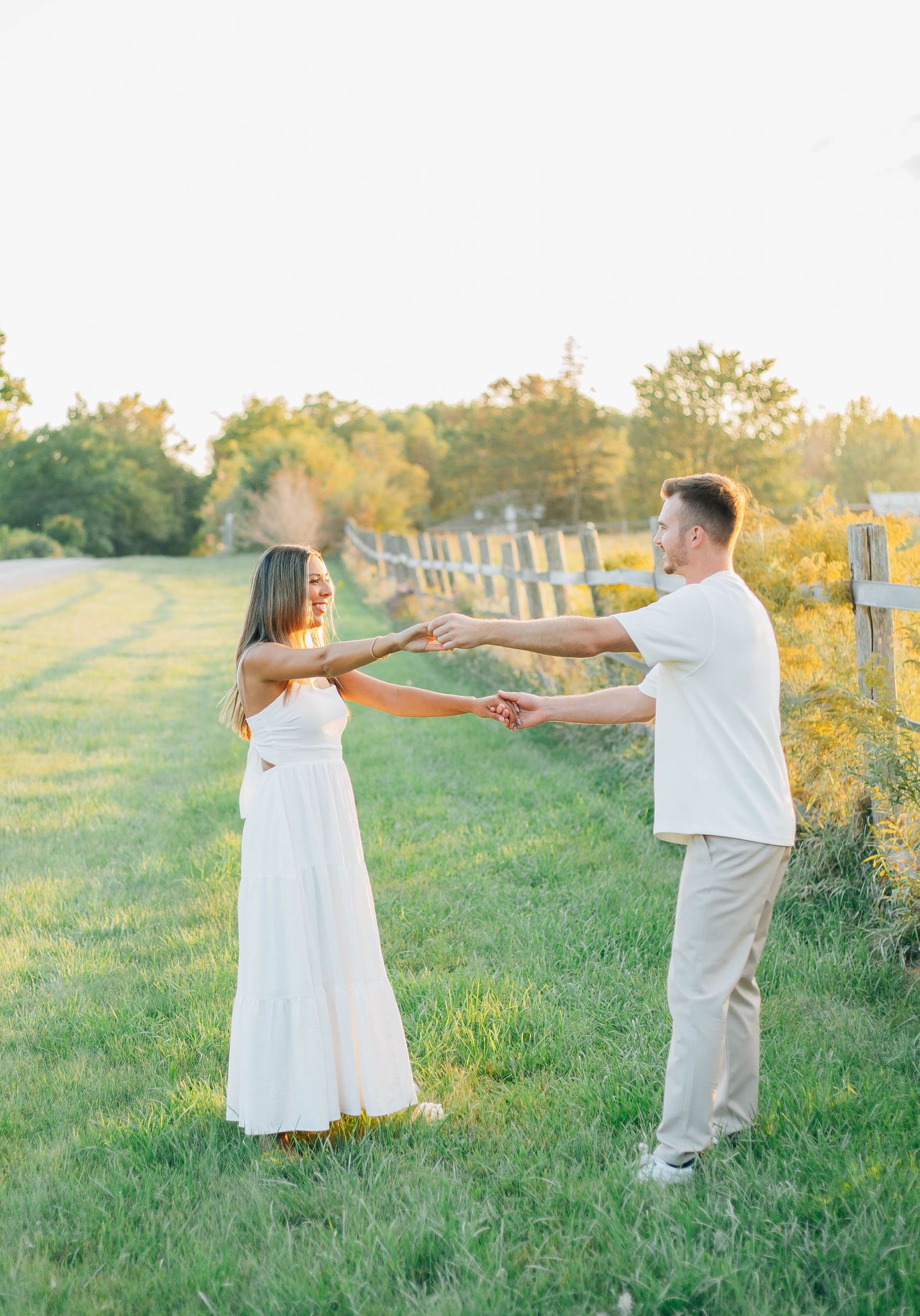 Couple holding hands and reaching toward each other in a golden sunlit green field at sunset.