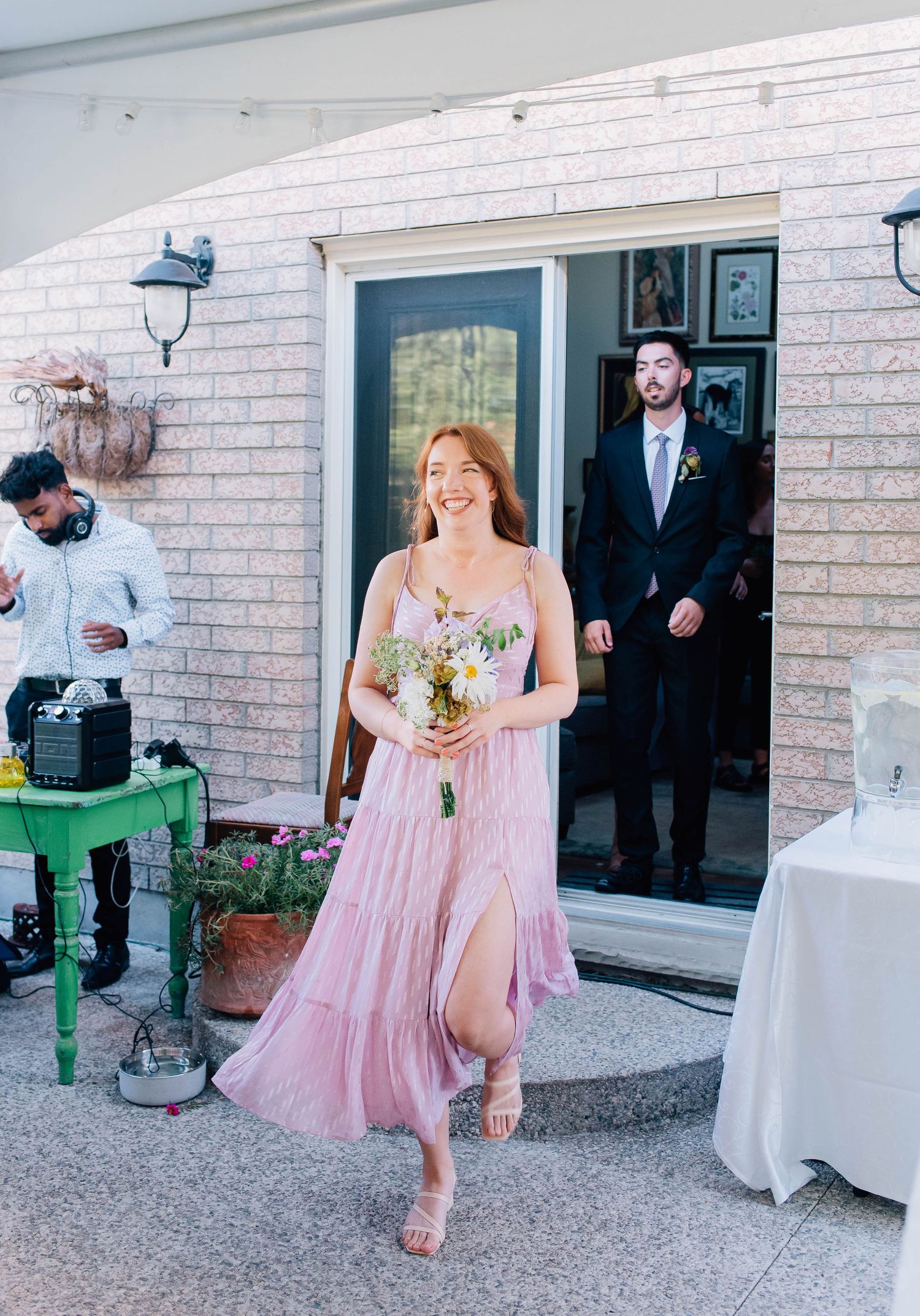 Bridesmaid in pink flowy dress walks down outdoor aisle carrying wildflower bouquet at sunny backyard wedding ceremony.