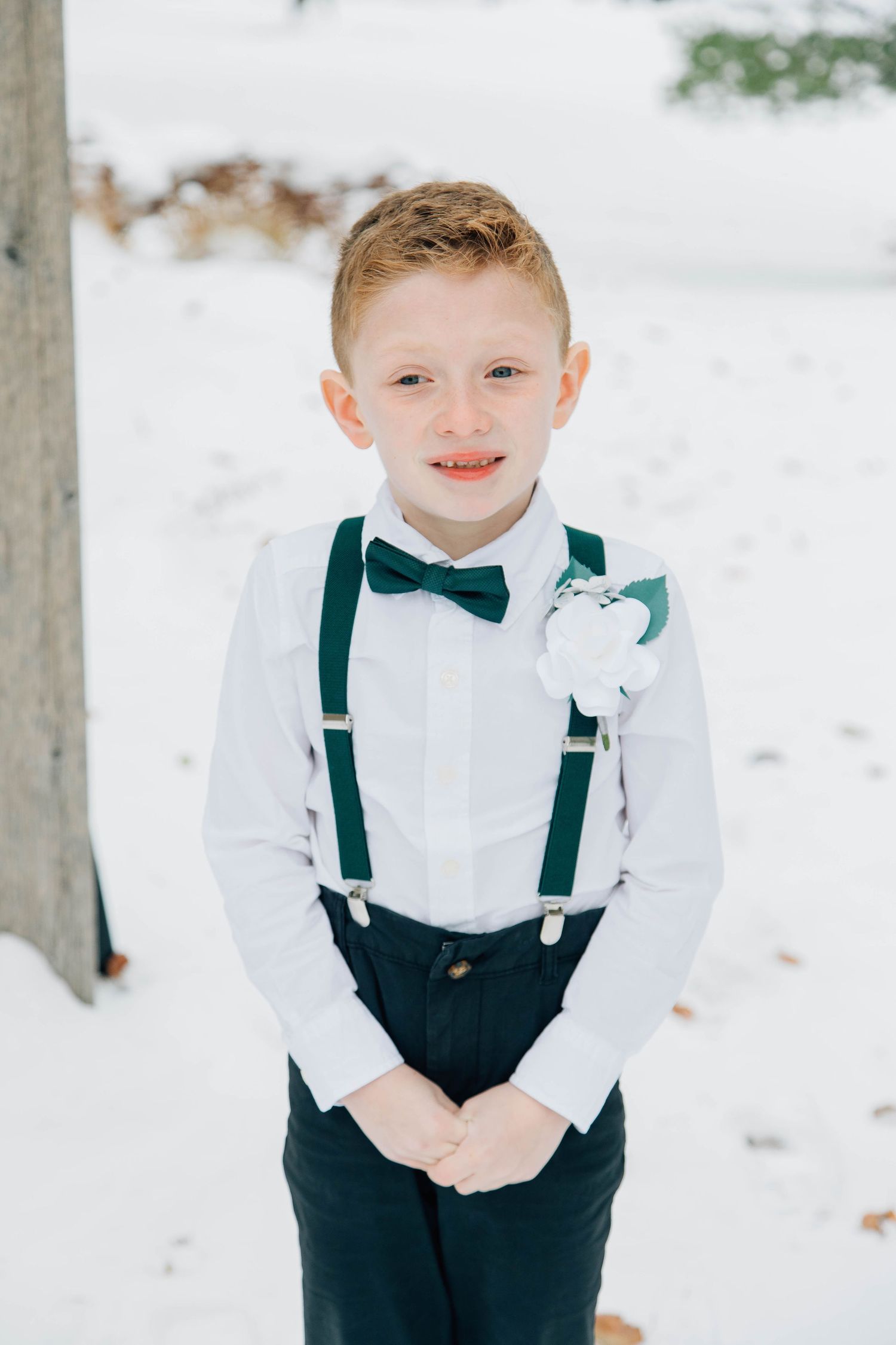 Young ring bearer in white shirt, green bow tie and suspenders smiles in snowy outdoor wedding.