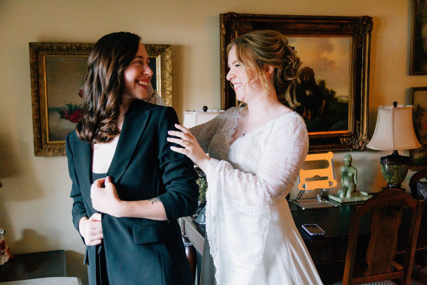 Bride in white dress smiles at partner in dark blazer during wedding preparation at home.