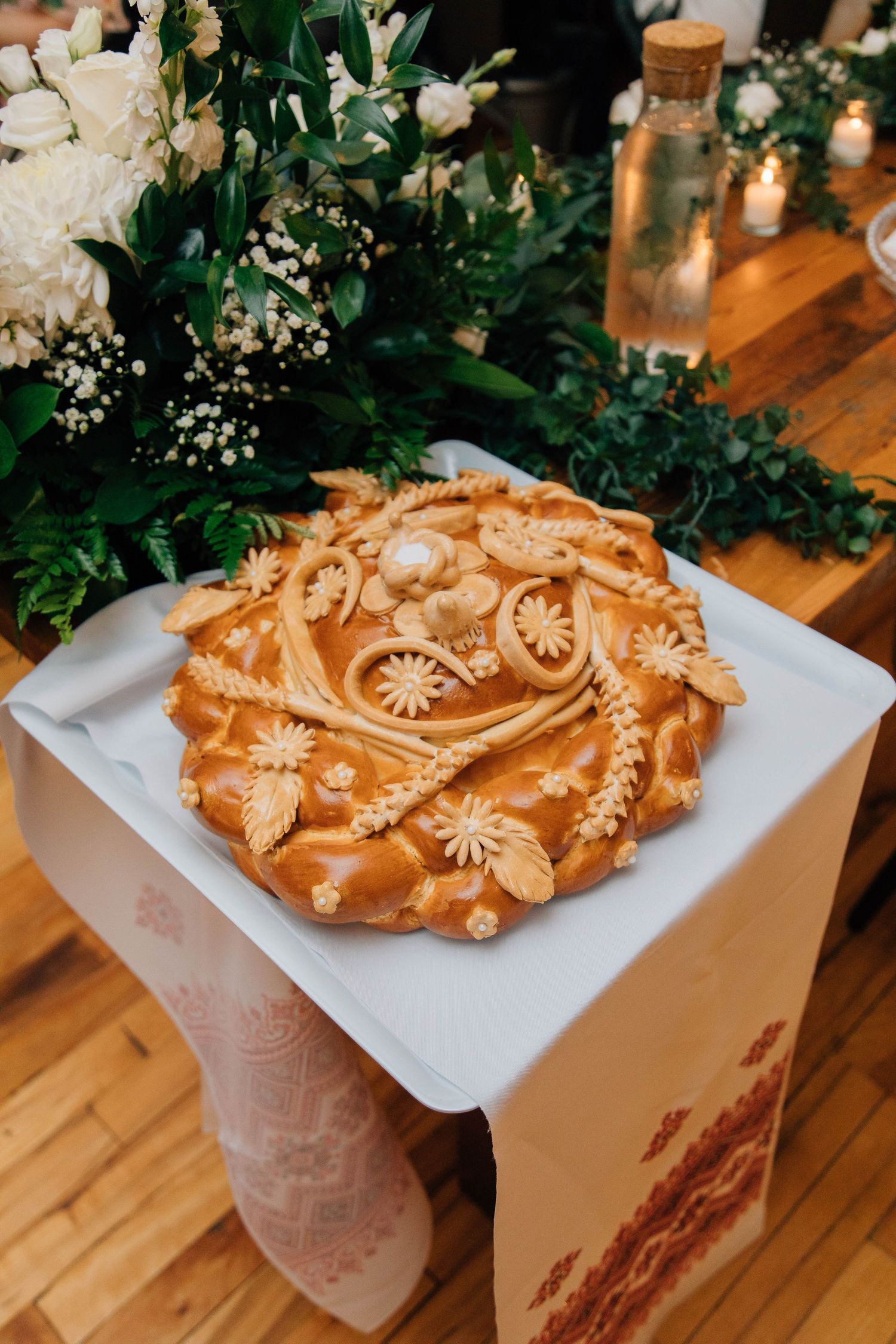 Ornate Ukrainian wedding bread korovai with intricate dough decorations on white cloth beside floral arrangements.