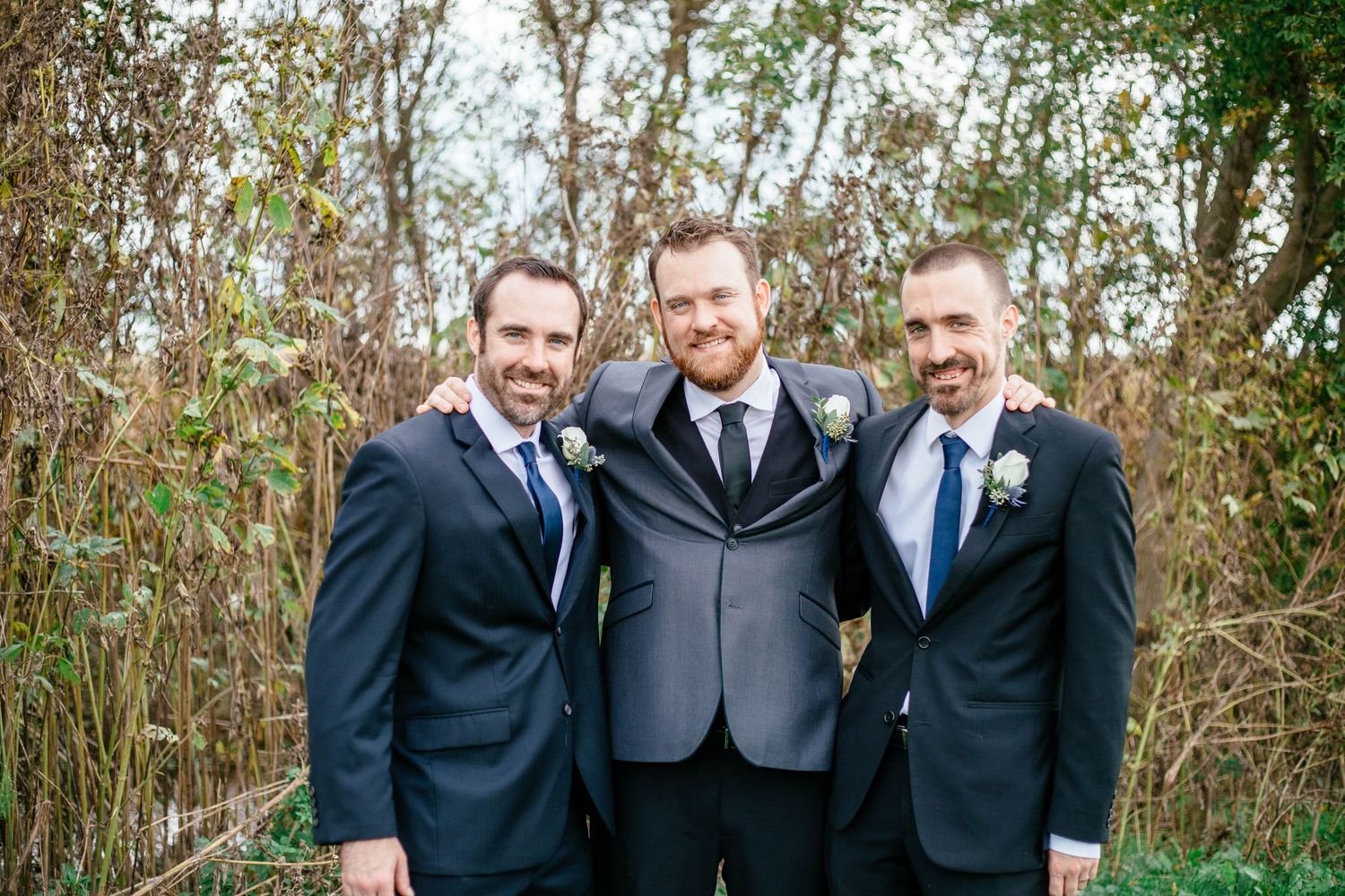 Groom in gray suit flanked by two groomsmen in navy suits smiling outdoors at fall wedding.