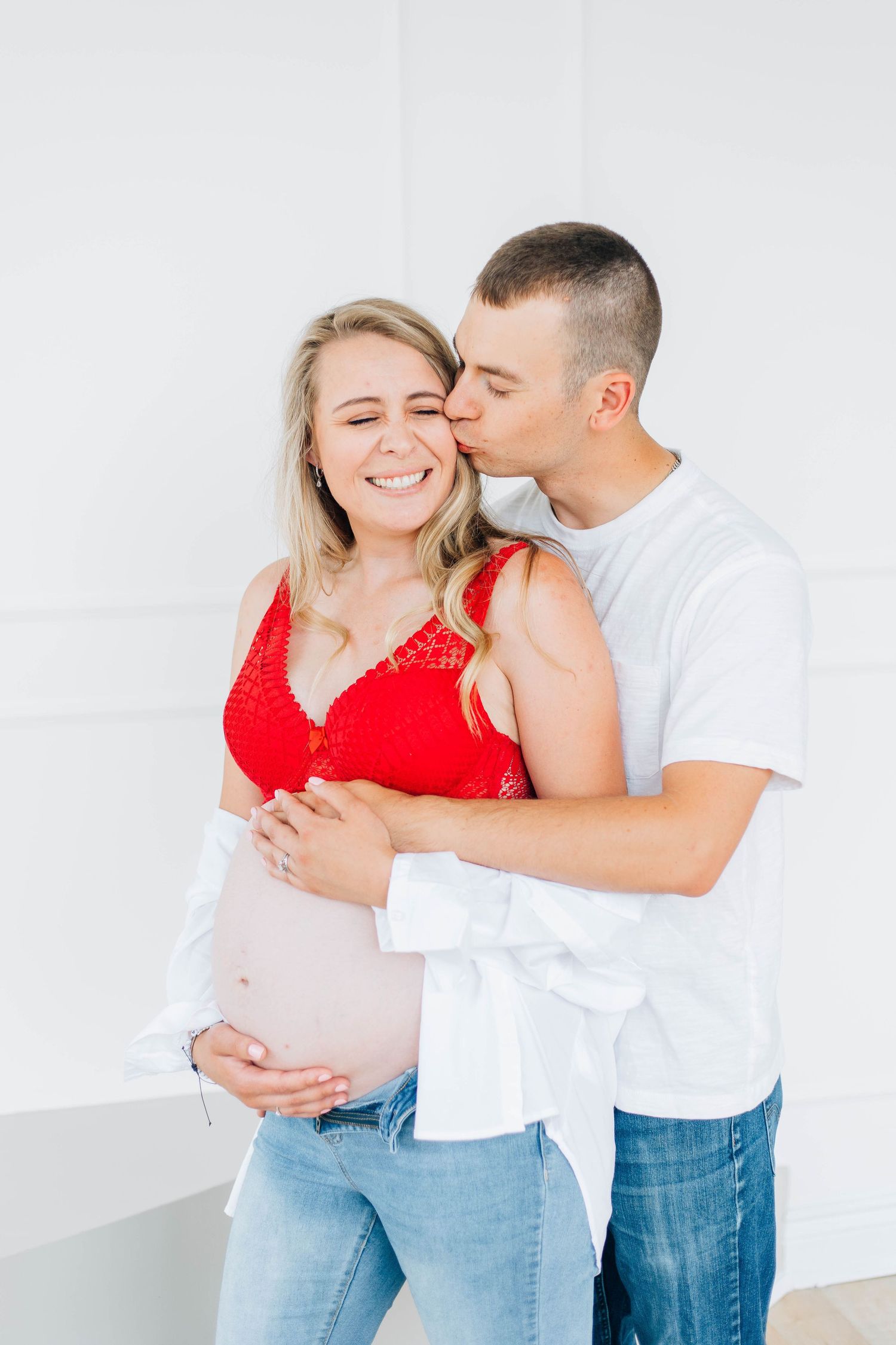 Man kissing smiling pregnant woman on cheek in red bralette and jeans.