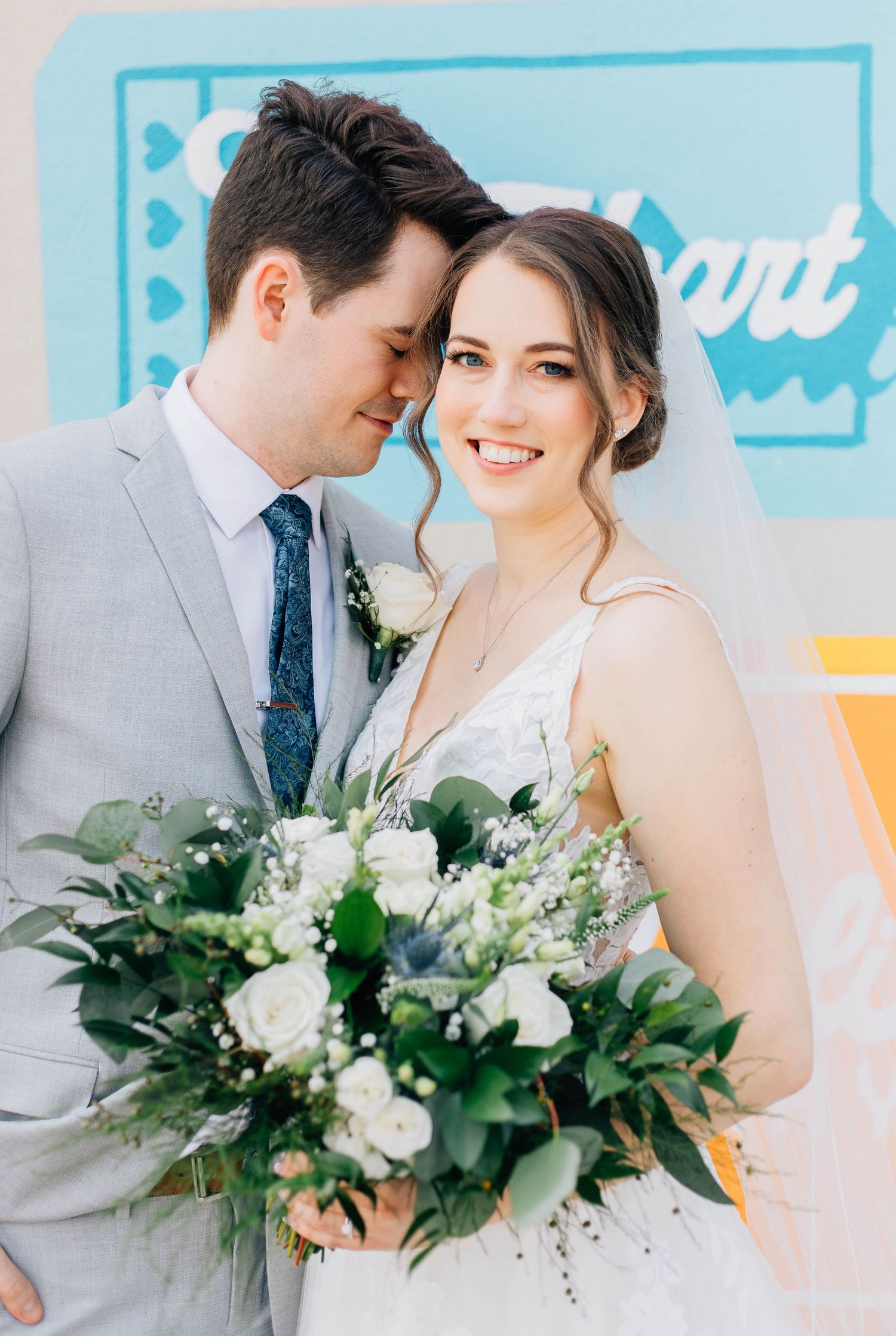 Bride holding white bouquet and groom in gray suit pose closely together at wedding.