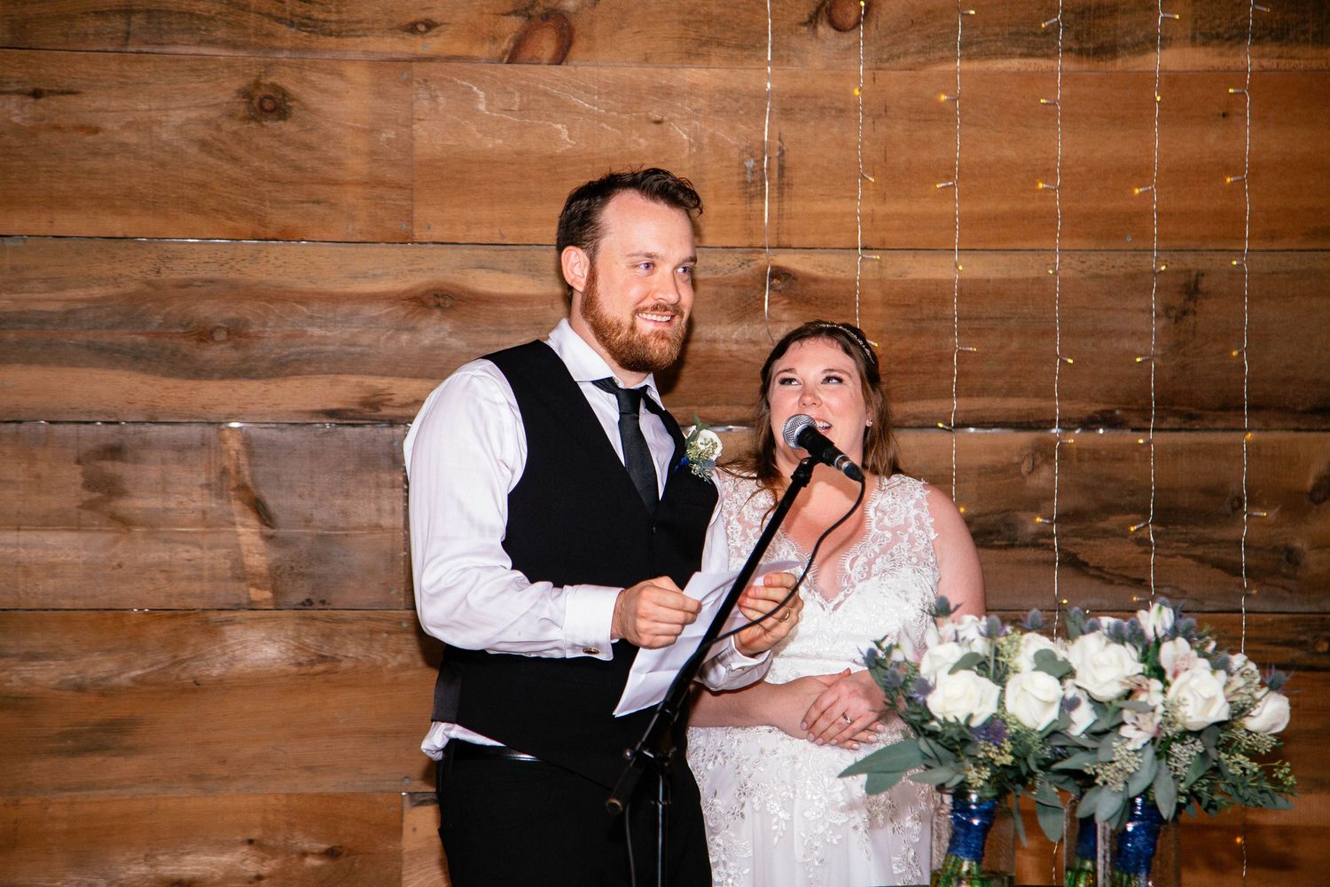 Bride and groom sharing a microphone during wedding reception speech in rustic barn venue.