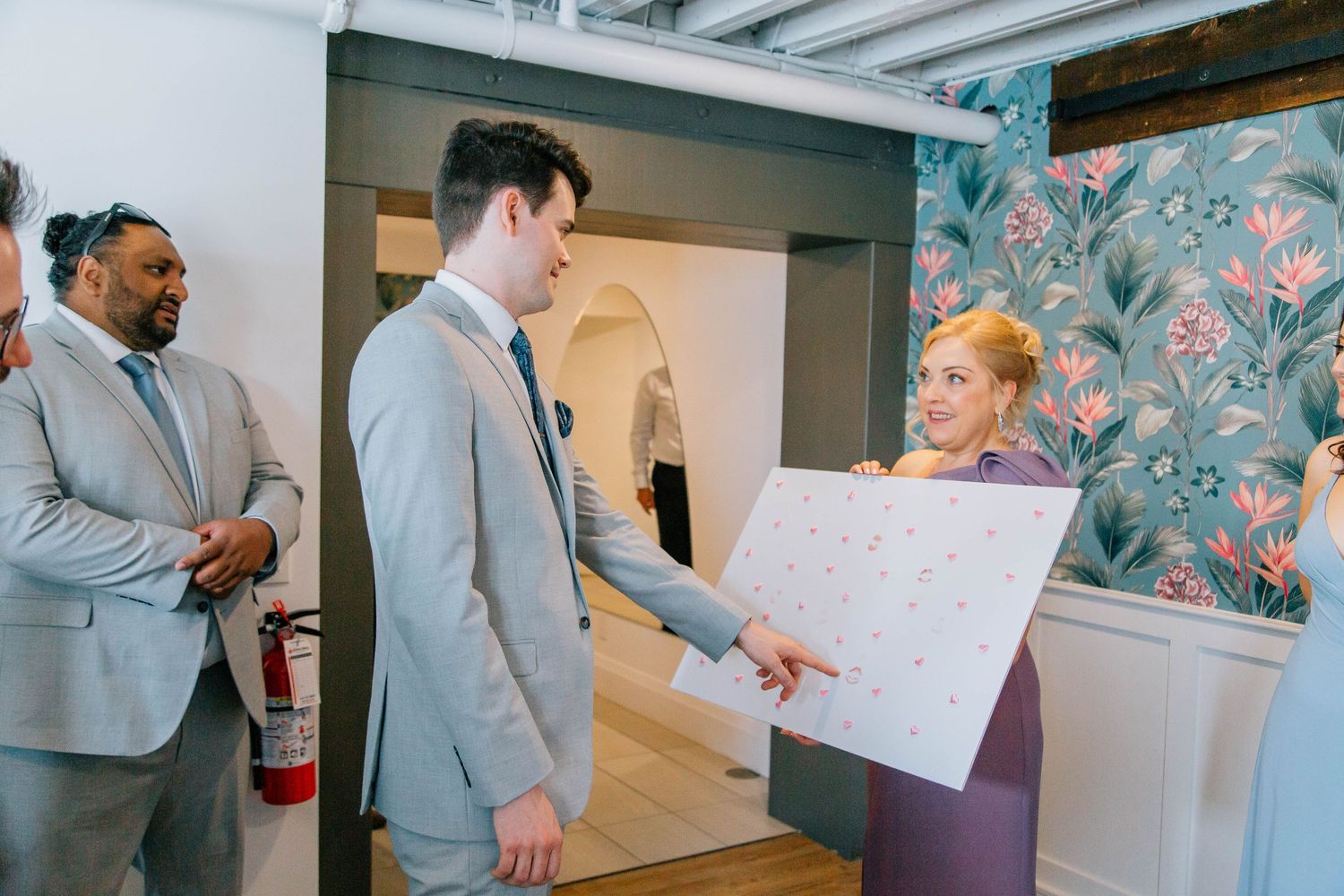 Groom in gray suit receives large decorative poster from bridesmaid in lavender dress at wedding venue.