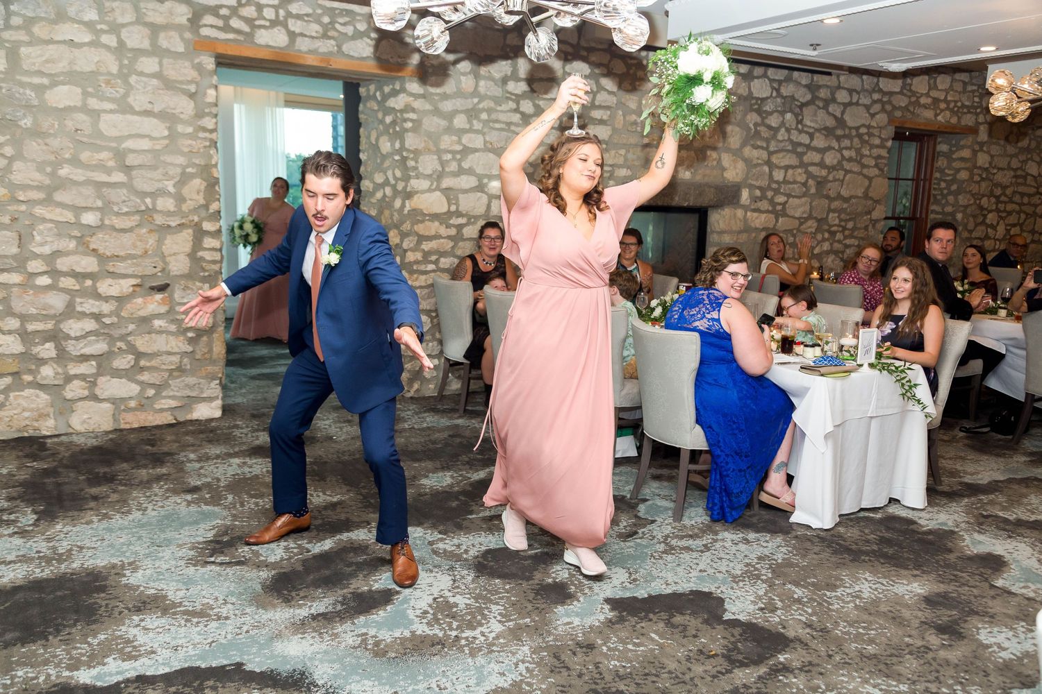 Bridesmaid in pink dress dances joyfully with groomsman at wedding reception in rustic stone venue.