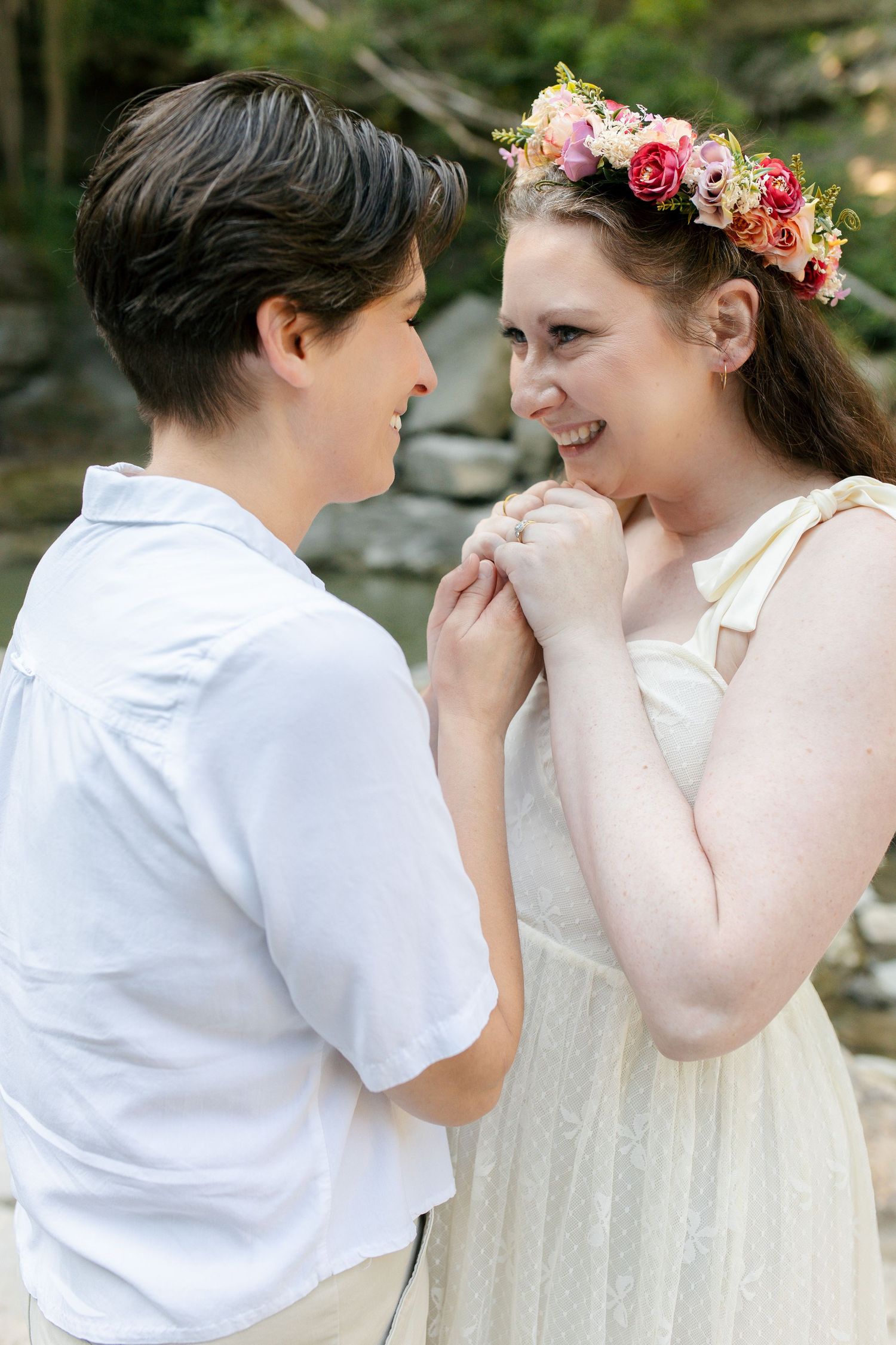 Two brides to be in white share a joyful moment, one wearing a floral crown, holding hands outdoors.