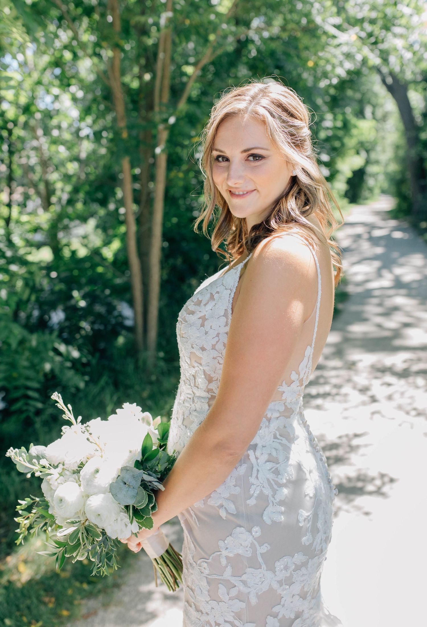 Bride in lace gown holding white bouquet on a sunlit garden path.