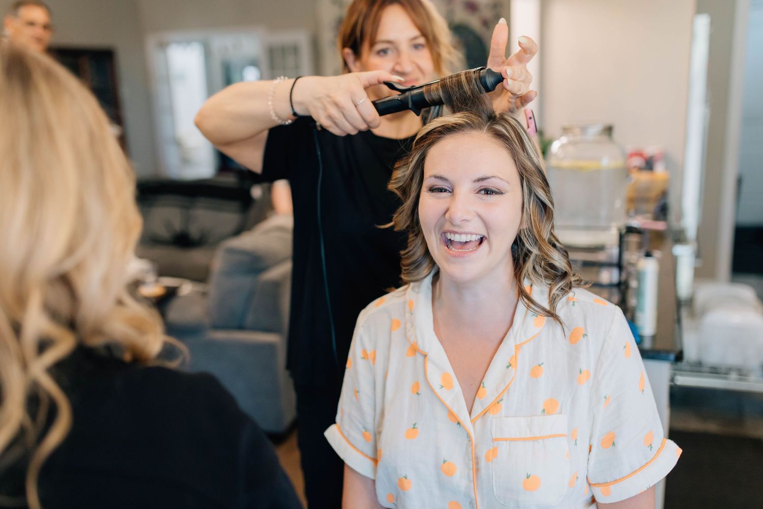 Bride laughing joyfully while getting her hair styled with a curling iron at a wedding salon.