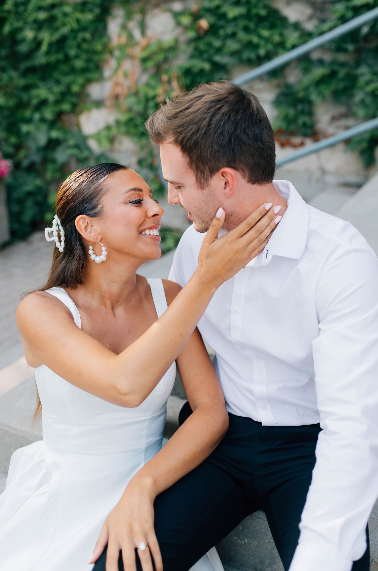 Couple gazes lovingly at each other while sitting on steps near ivy-covered stone wall.