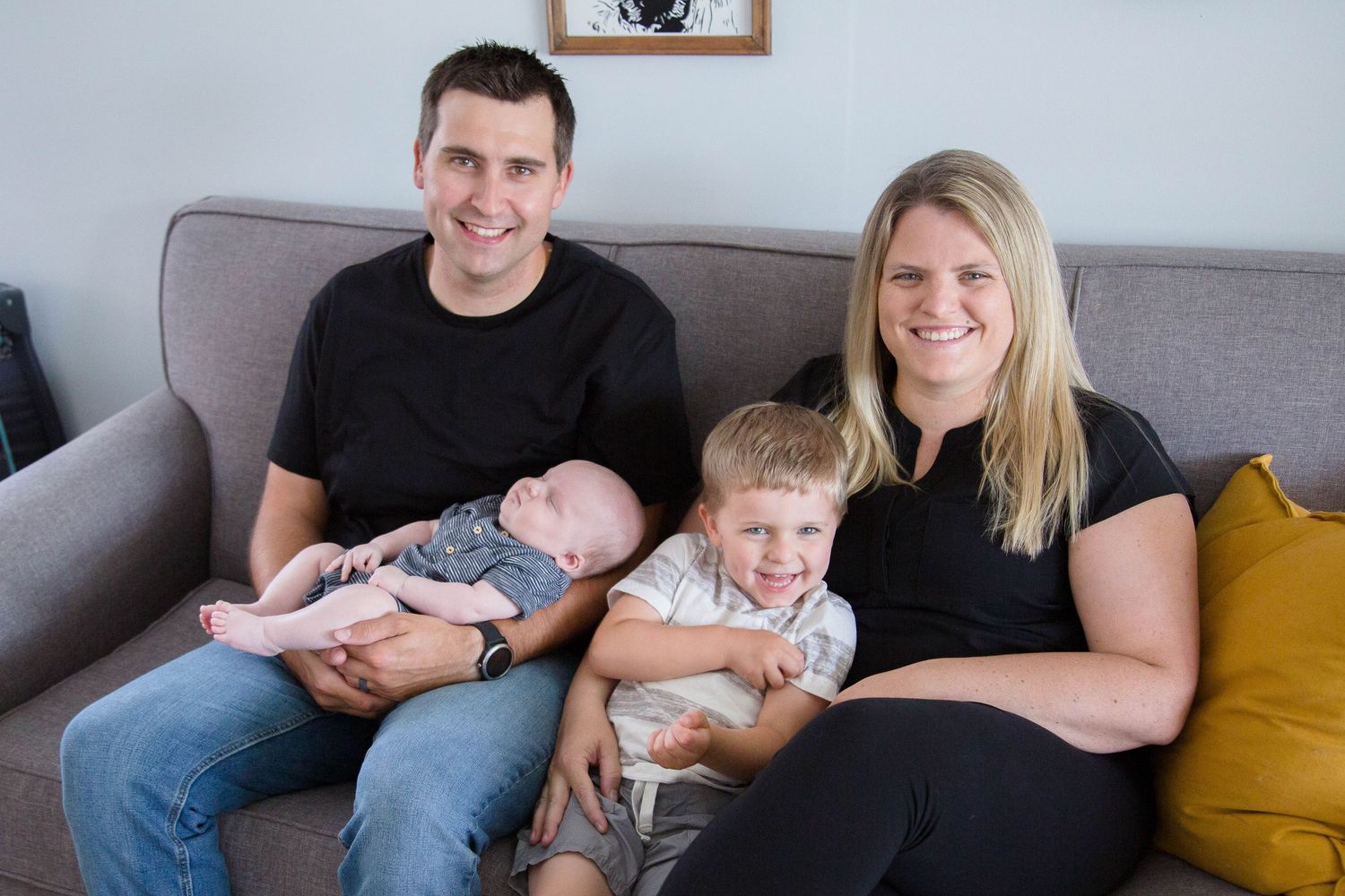 Family portrait of parents with newborn baby and young toddler son sitting together on gray couch at home.