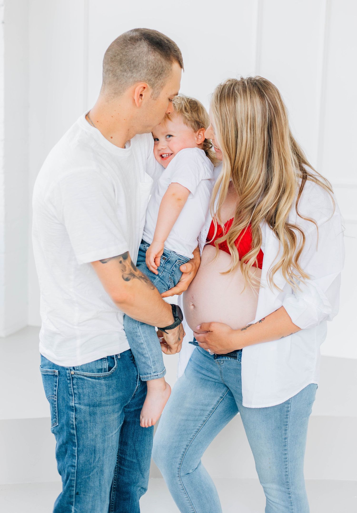 Joyful family maternity photo with tattooed dad and pregnant mom in red bralette kissing laughing toddler.