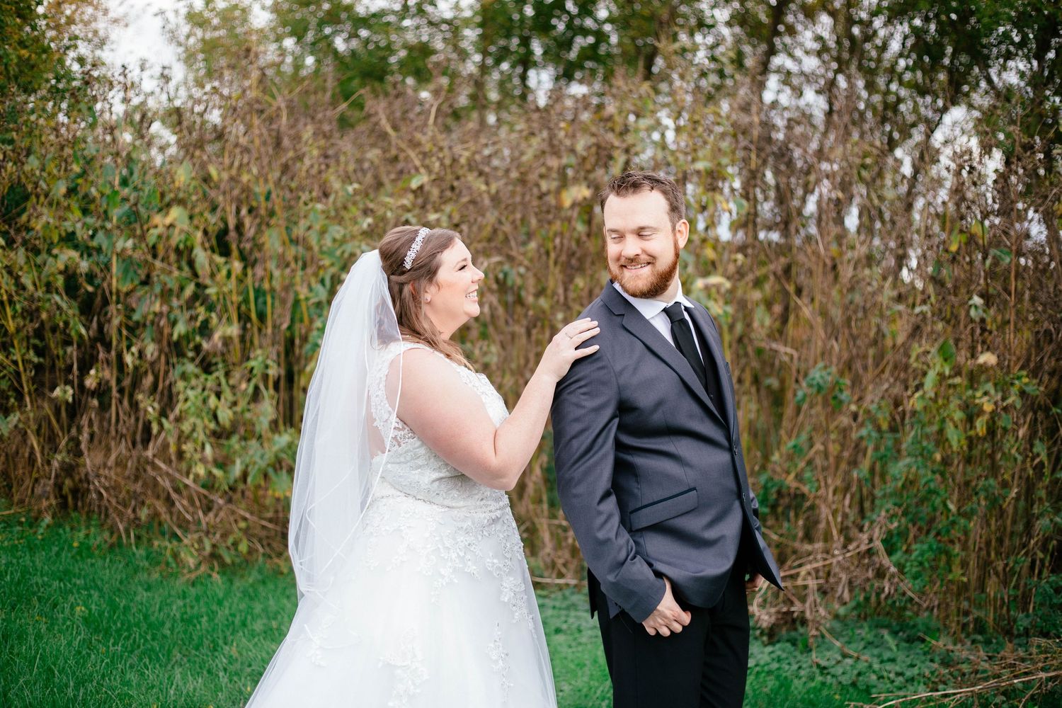 Bride in lace gown and veil touching groom's shoulder during romantic first look outdoors.