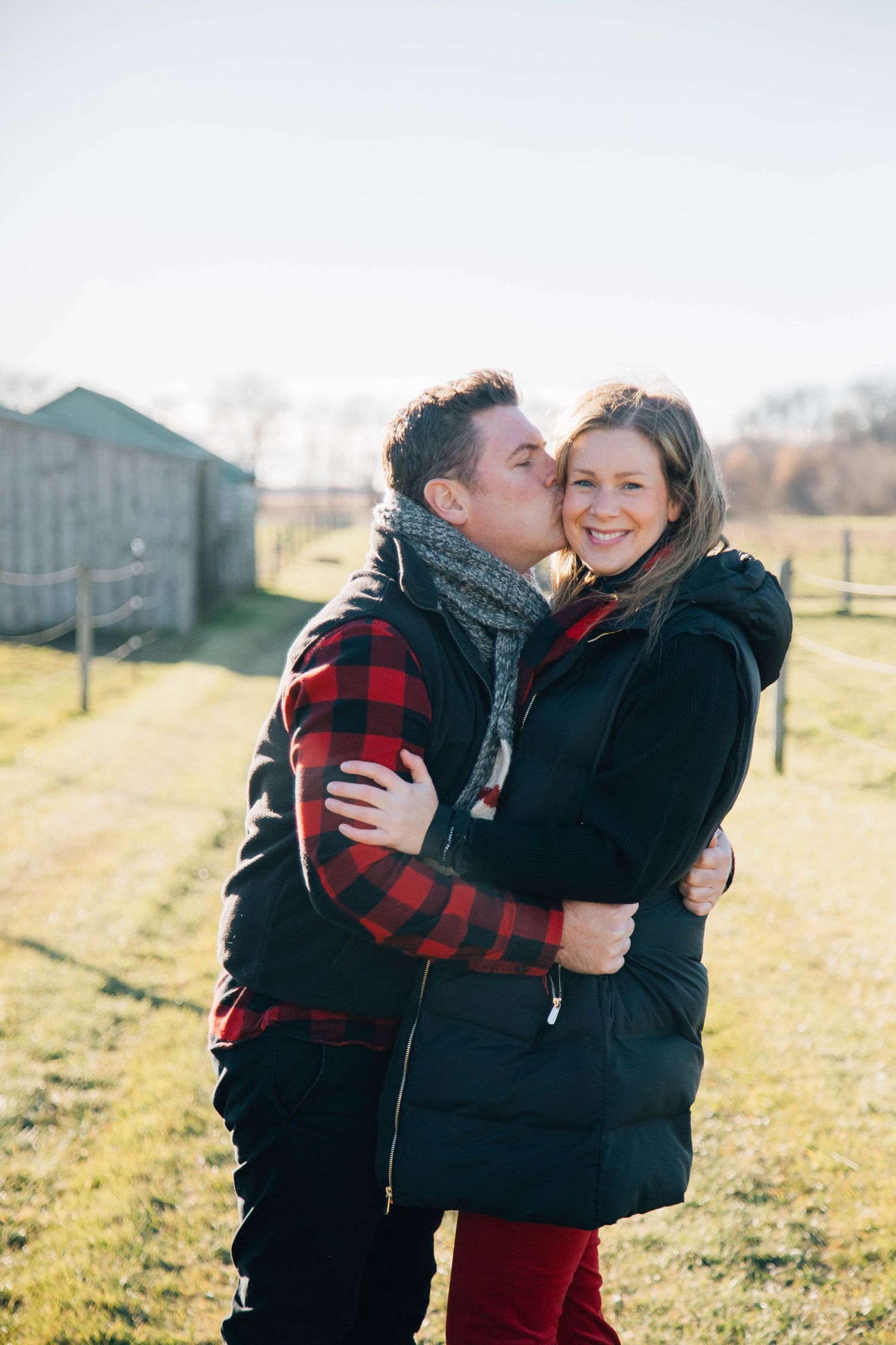 Couple embracing outdoors on a sunny farm, man kissing woman on the cheek while she smiles.