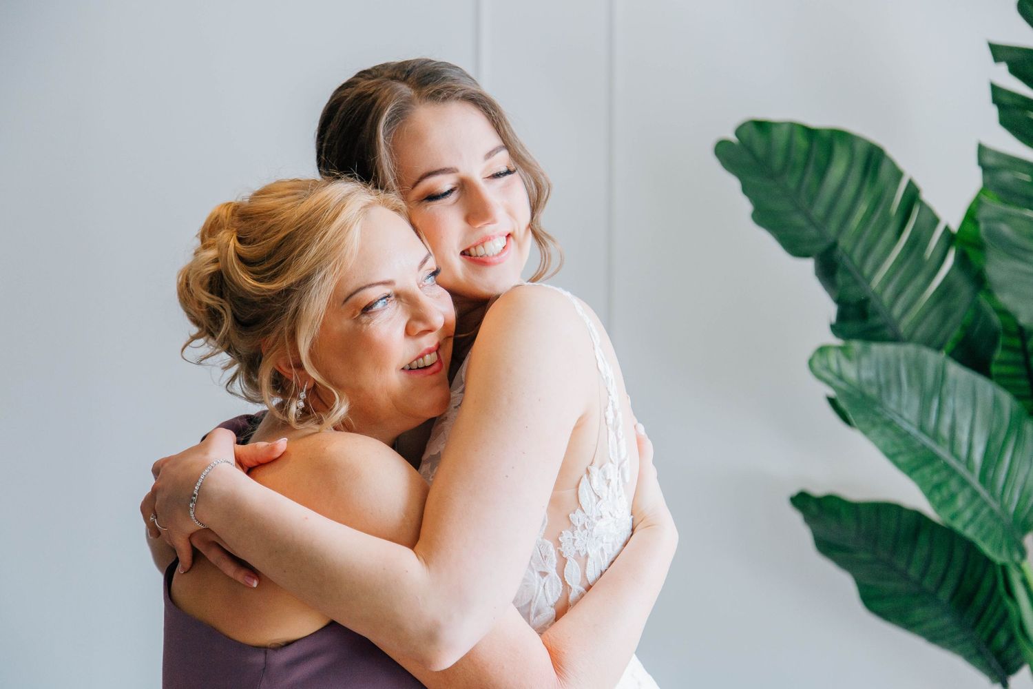 Bride in white lace gown shares a warm embrace with a bridesmaid in a lavender dress.