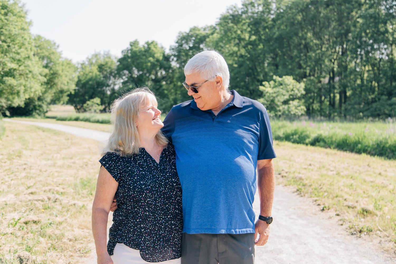 Older couple smiling at each other warmly while standing together on a sunny outdoor path with green trees.