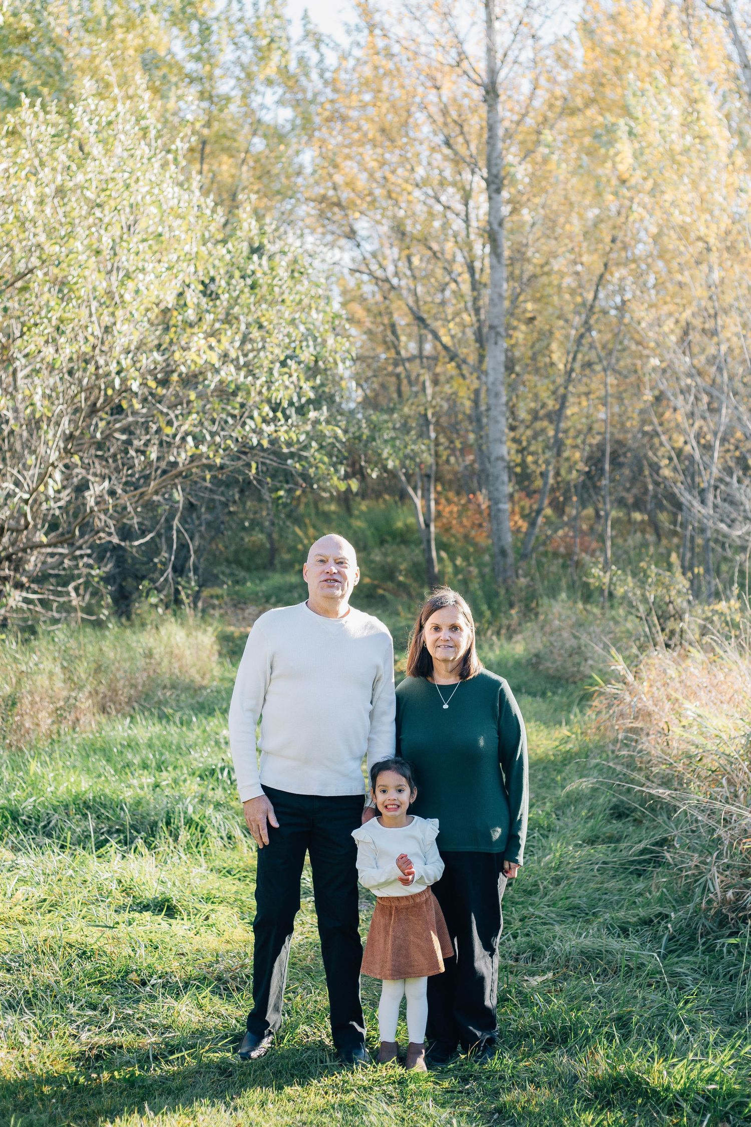 Grandparents and young granddaughter posing together in a sunny autumn outdoor setting.