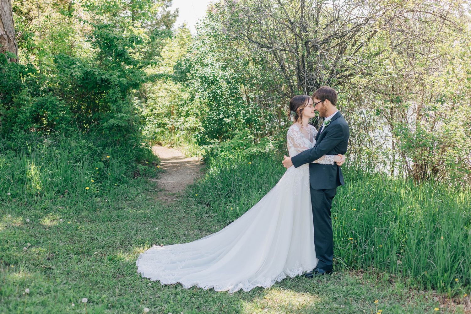 Bride and groom sharing a romantic embrace on a lush green garden path surrounded by trees.