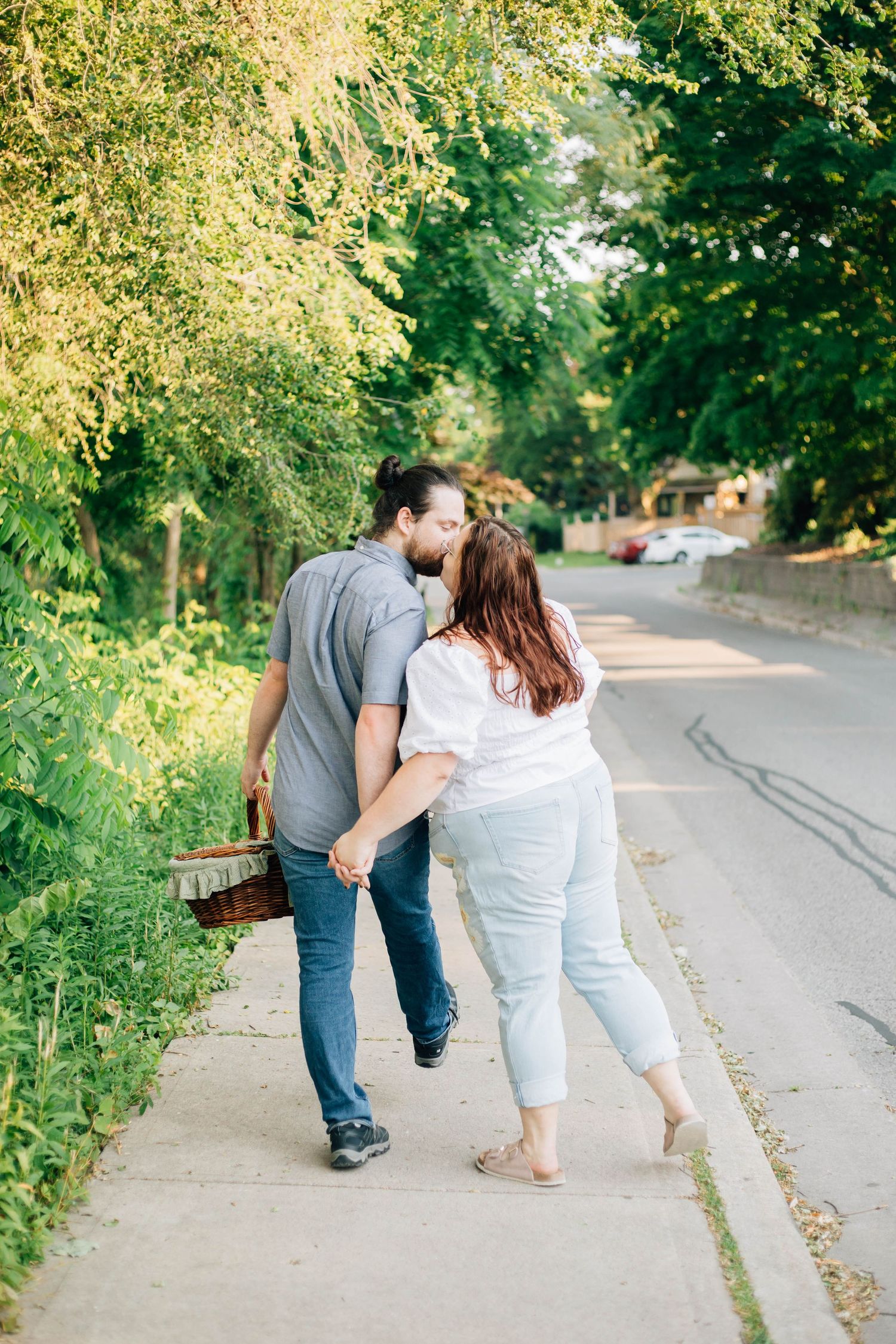 Couple stealing a kiss while walking on a sunny tree-lined sidewalk with a picnic basket.