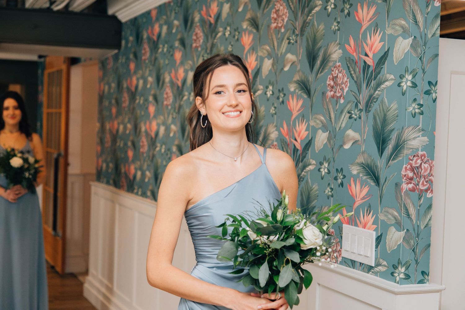 Smiling bridesmaid in blue dress holding bouquet before floral wallpaper.
