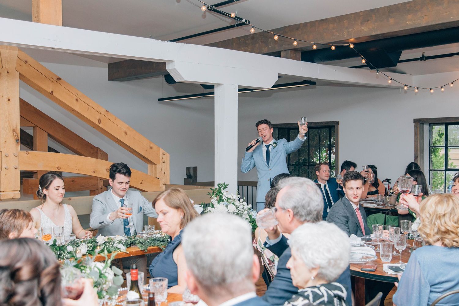 Best man giving a toast speech at rustic barn wedding reception with string lights overhead.