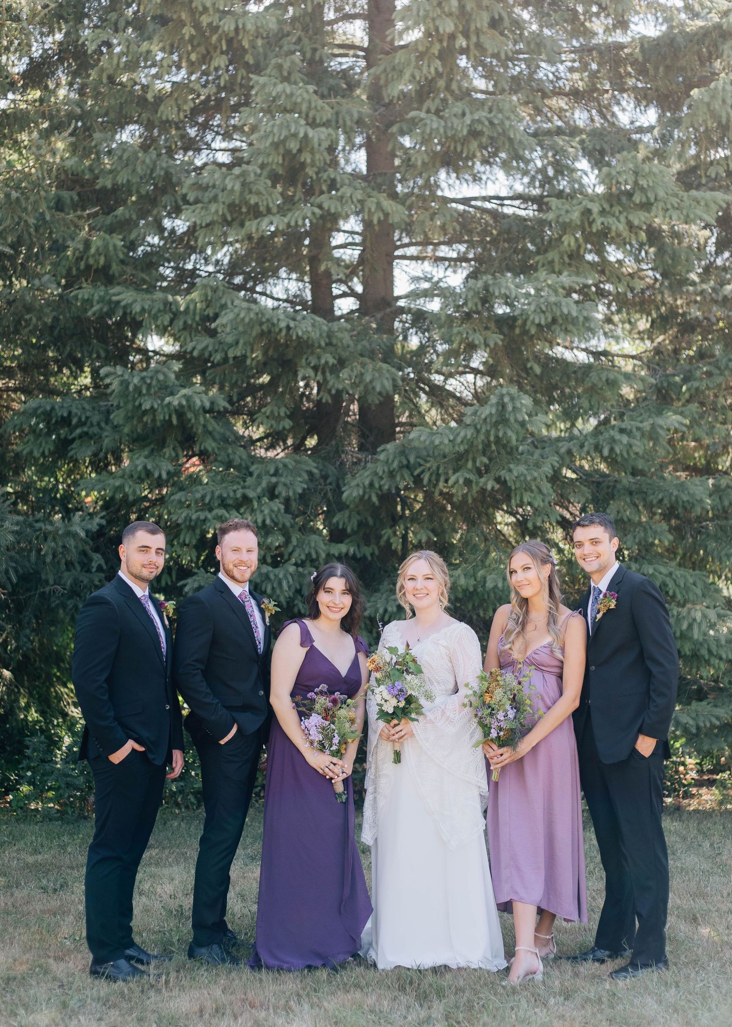 Wedding party of six posing formally in front of tall evergreen tree with bridesmaids in purple gowns.