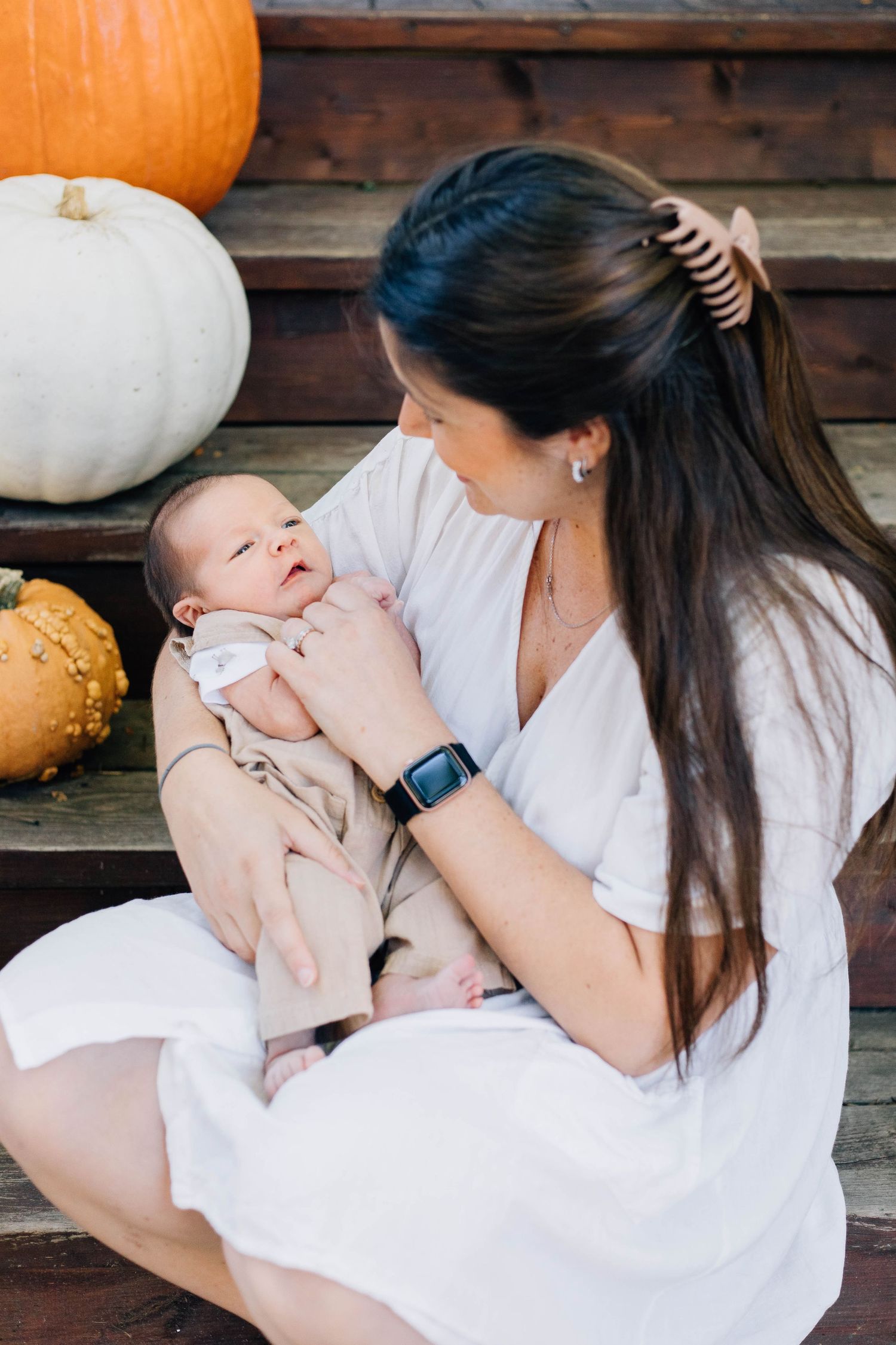 Dark-haired mother in white dress sitting on fall porch steps holding newborn baby, surrounded by orange and white pumpkins.