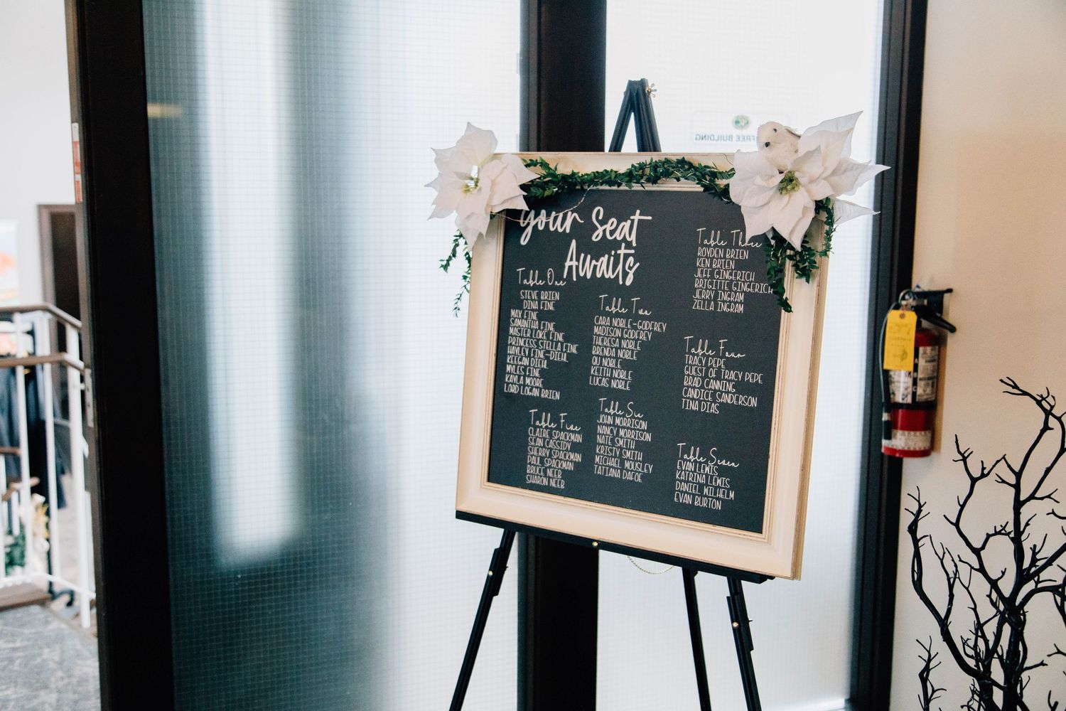 Wedding seating chart displayed on a chalkboard easel decorated with white poinsettias and greenery.