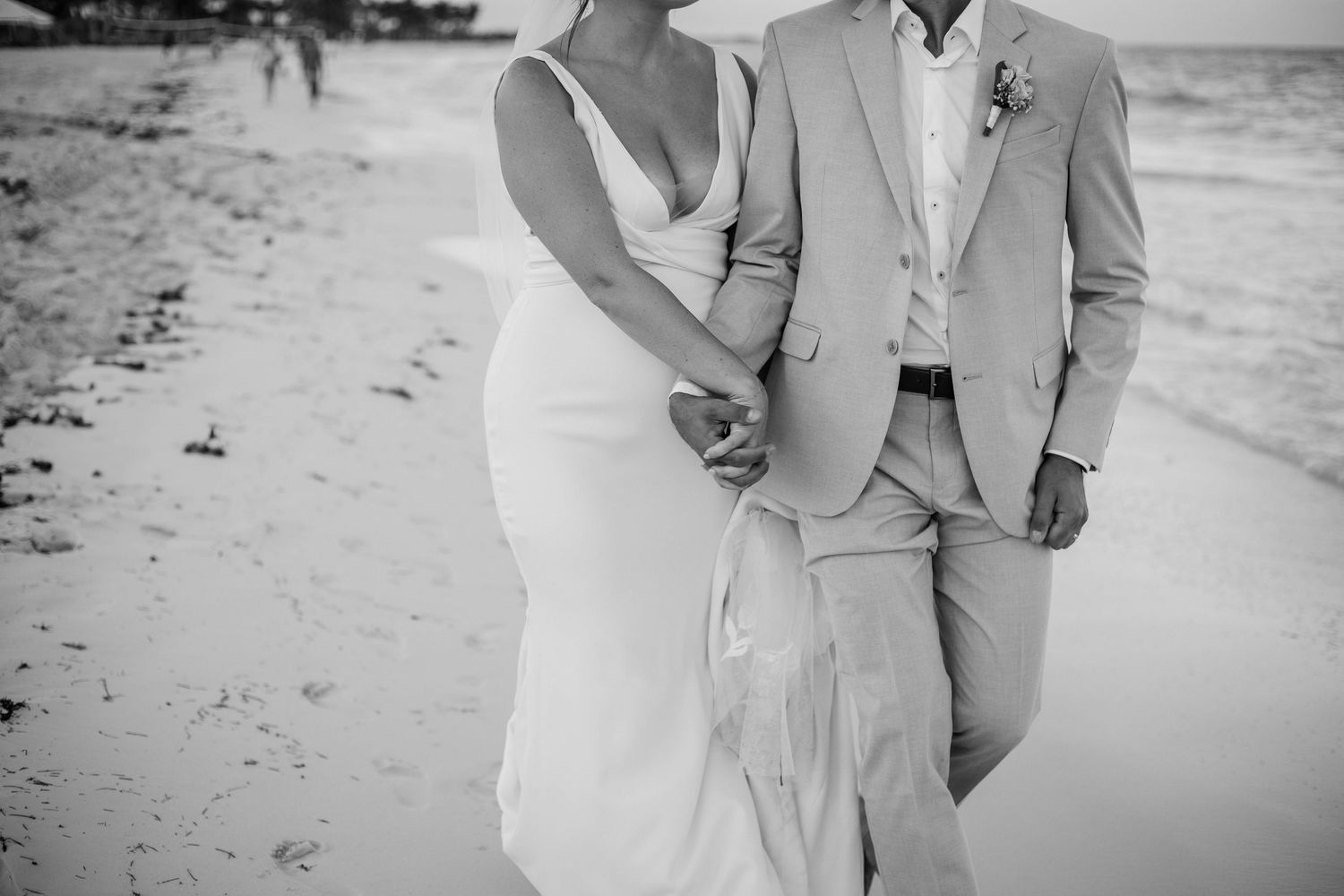 Black and white photo of a bride and groom holding hands and walking along a sandy beach.