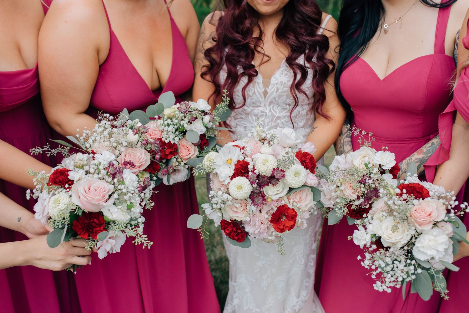Bride in lace gown with bridesmaids in pink dresses holding elegant red and white floral bouquets.