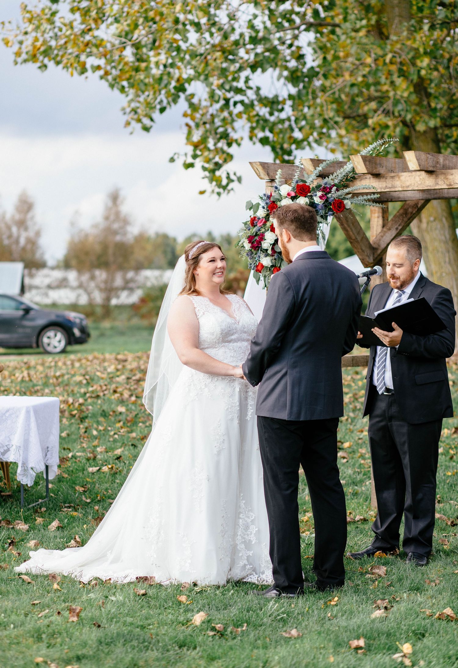 Bride in lace gown holds hands with groom at outdoor fall wedding ceremony with red florals.