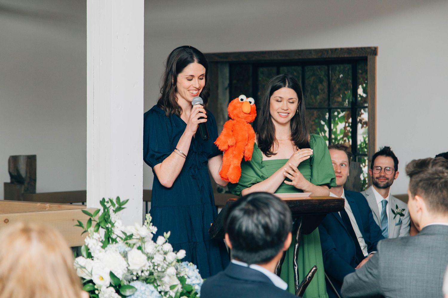 Two bridesmaids entertaining wedding guests with an Elmo puppet during reception speeches.