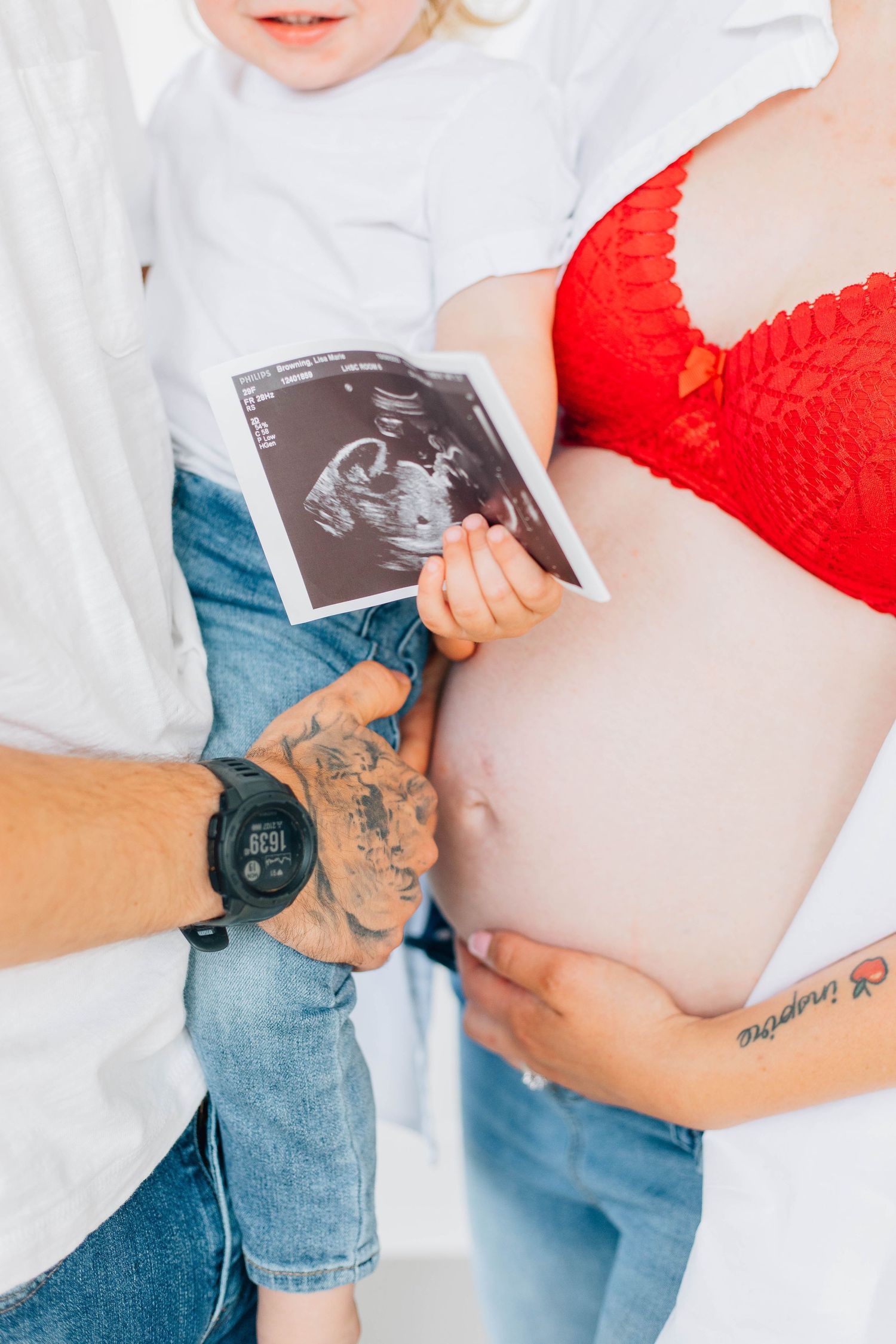 Close-up of pregnant belly with tattooed dad and toddler holding ultrasound photo during maternity session.