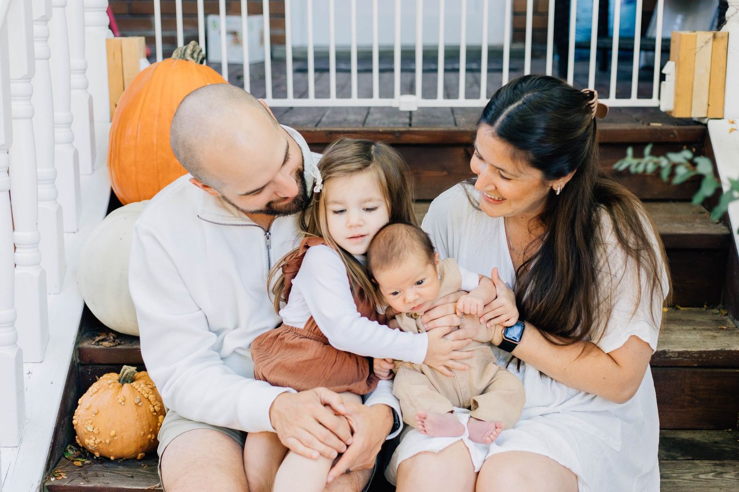 Happy family portrait on autumn decorated steps, parents and toddler girl lovingly gazing at newborn baby surrounded by pumpkins.