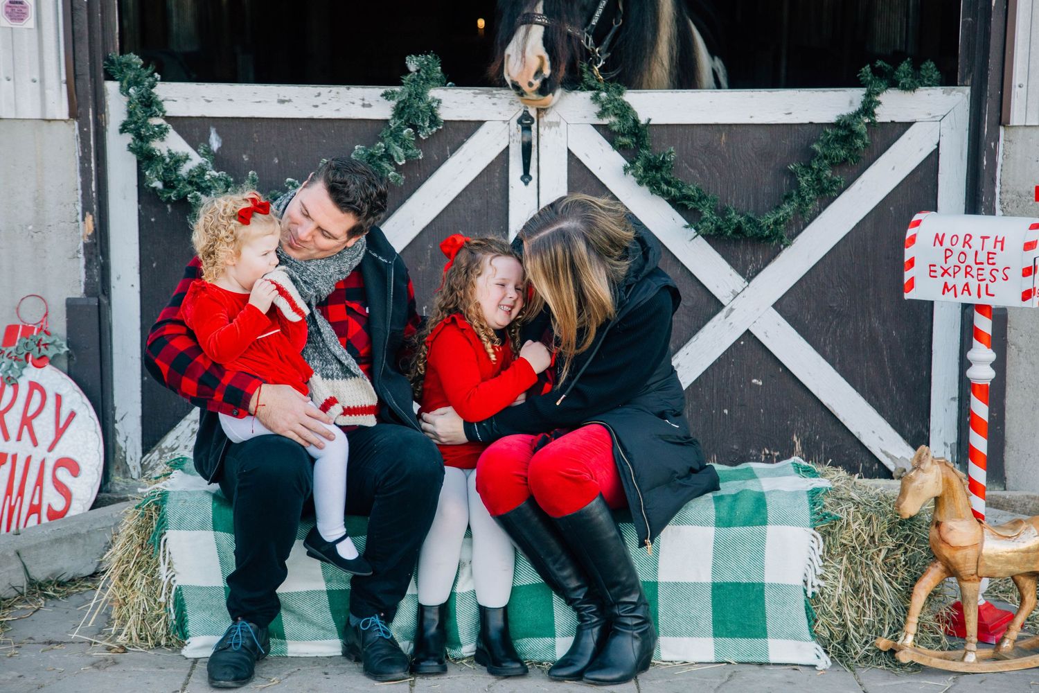 Family of four in red Christmas outfits sitting on hay bales in front of a decorated barn.
